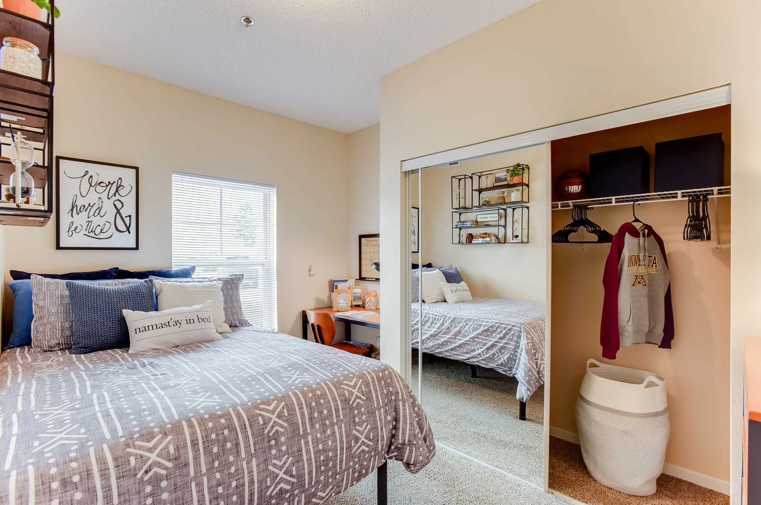 A bedroom with two beds, one in the foreground and one in a mirrored closet. The beds have gray and white bedding and pillows, one of which has the text 'namastây in bed.' There is a window on the left wall, and decorative wall art saying 'Work hard & be nice.' The closet has an open section with hanging clothes, and a basketball and two TV monitors on a wire shelf.