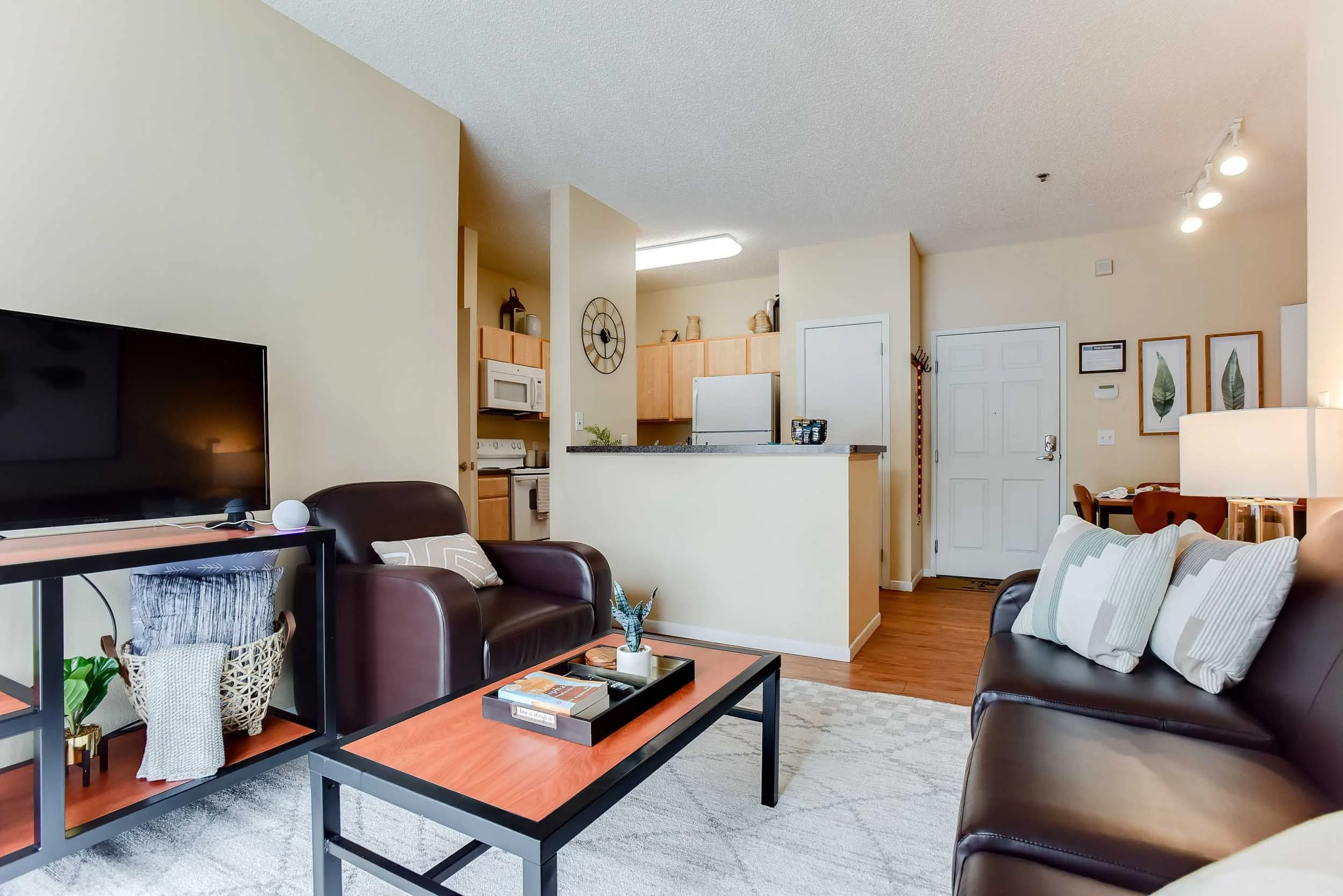 Living room with brown leather sofas, a black and orange coffee table, a TV, and a side table with plants and decorative items. Open kitchen visible in the background with wooden cabinets and a refrigerator.