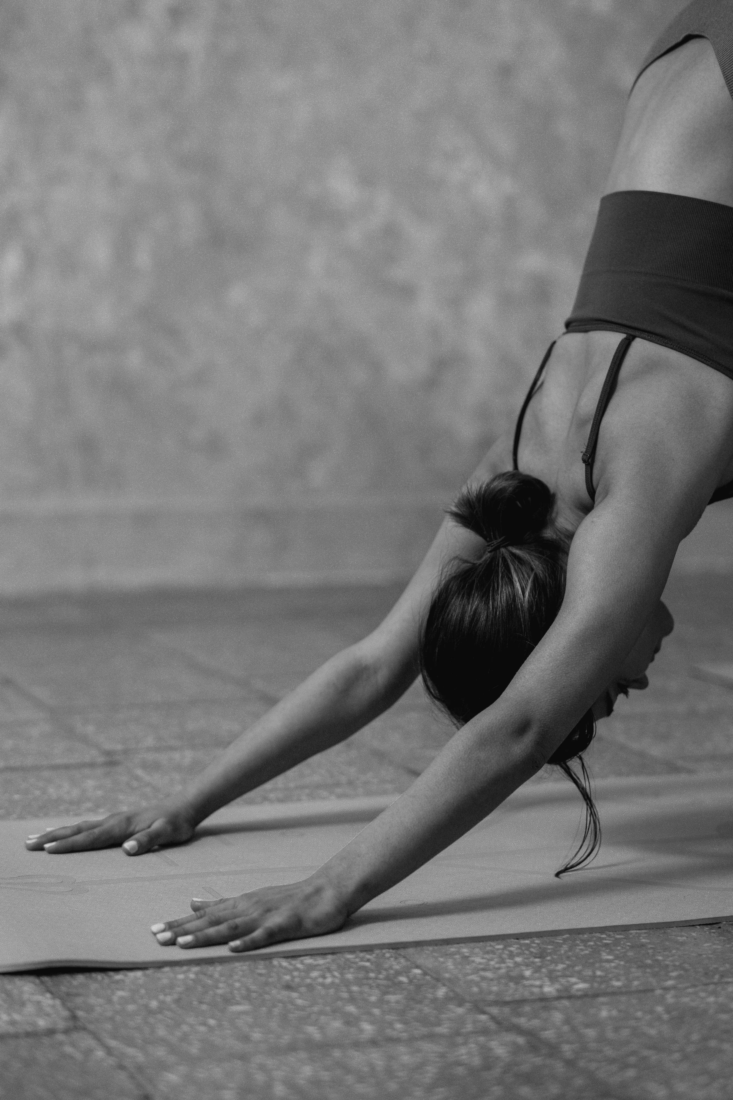 A woman performing yoga outdoors on a yoga mat, with her body bent forward and her head resting on her legs, on a paved surface.