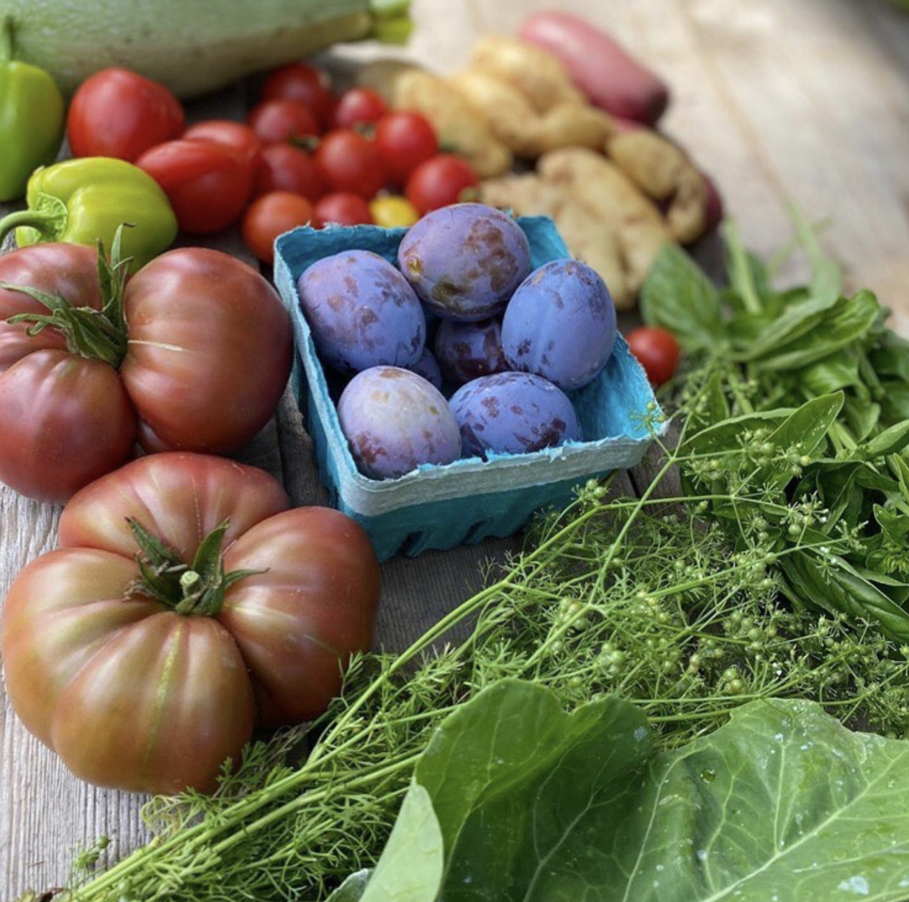 A variety of fresh vegetables and fruits including heirloom tomatoes, grapes, cherry tomatoes, ginger, and assorted leafy greens displayed on a wooden surface.