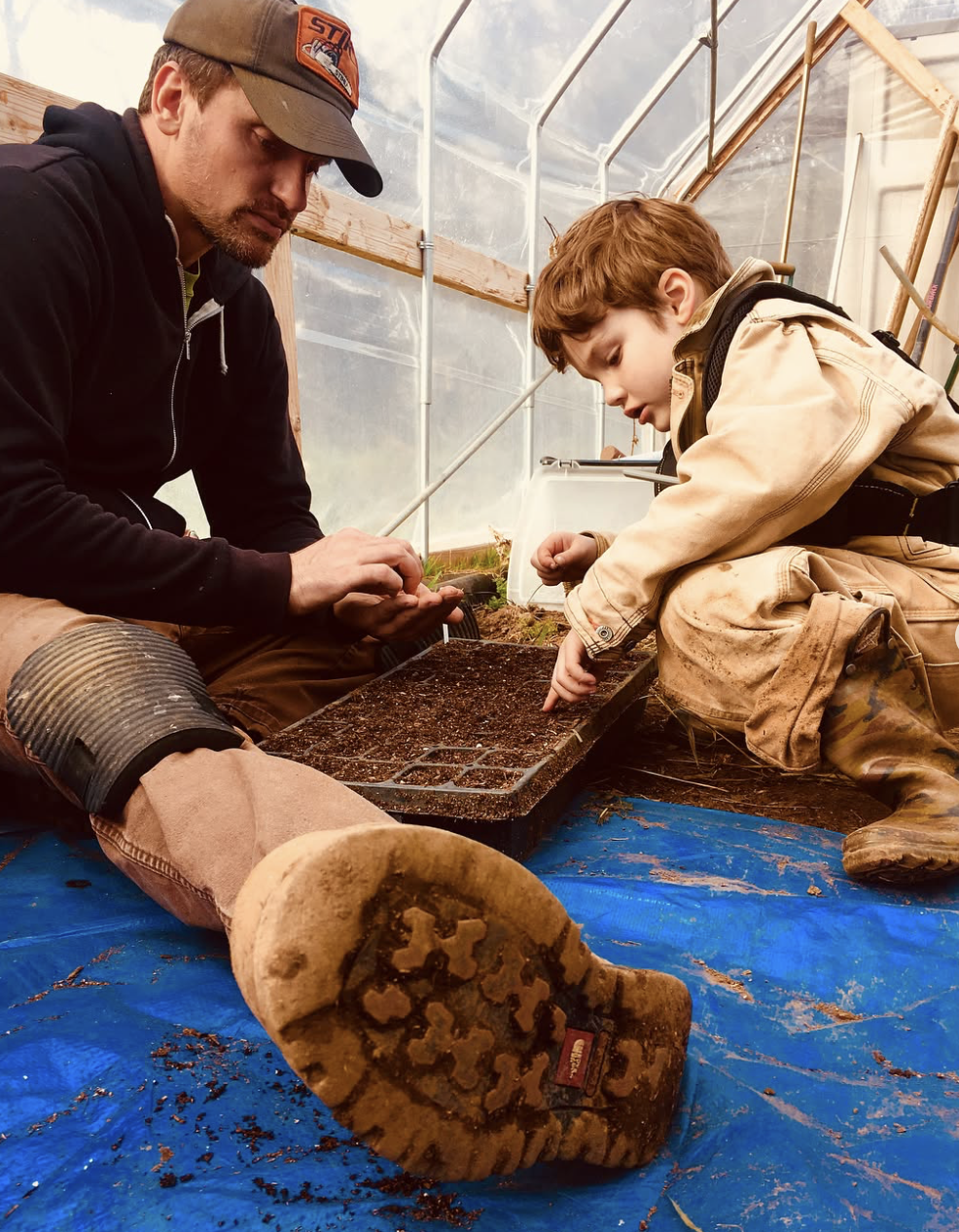 A man and a young boy planting seeds in a tray inside a greenhouse. The man is wearing a black hoodie and a cap, and the boy is in dirty work clothes, kneeling on a blue tarp.