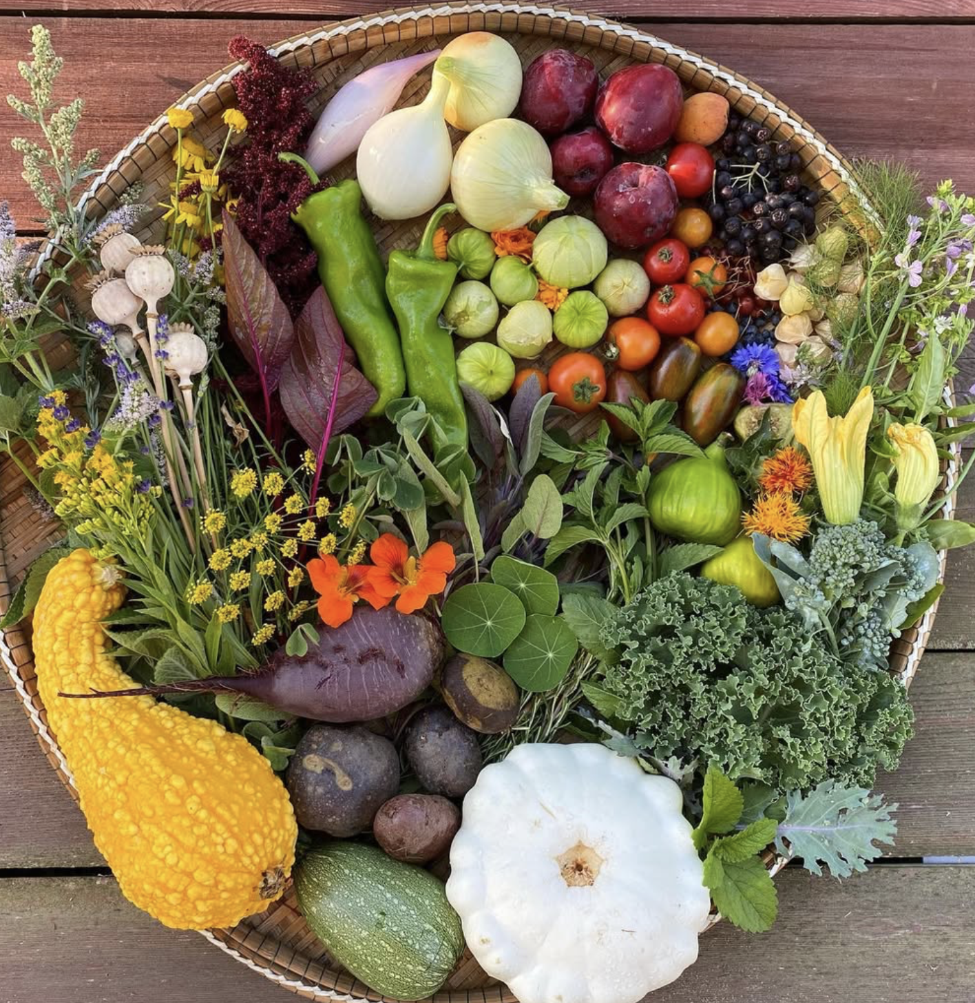 A wicker basket filled with fresh vegetables, fruits, and flowers on a wooden surface, including squash, tomatoes, eggplants, peppers, berries, and various colorful flowers.