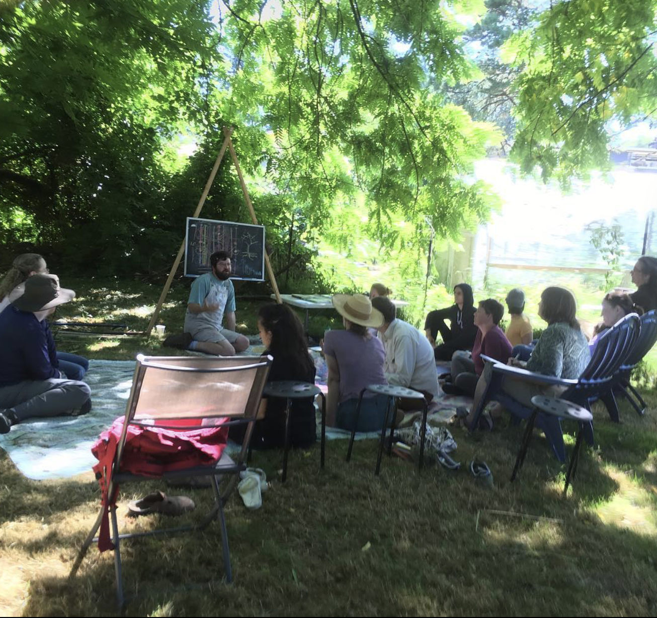 A group of people sitting on a blanket and chairs outdoors under large leafy trees, attending a presentation or class led by a man seated on the ground near a blackboard with drawings. Sunlight filters through the branches, creating a shaded setting 