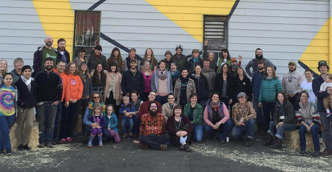 A large group of people of various ages assembled outdoors in front of a colorful building, posing for a group photo.