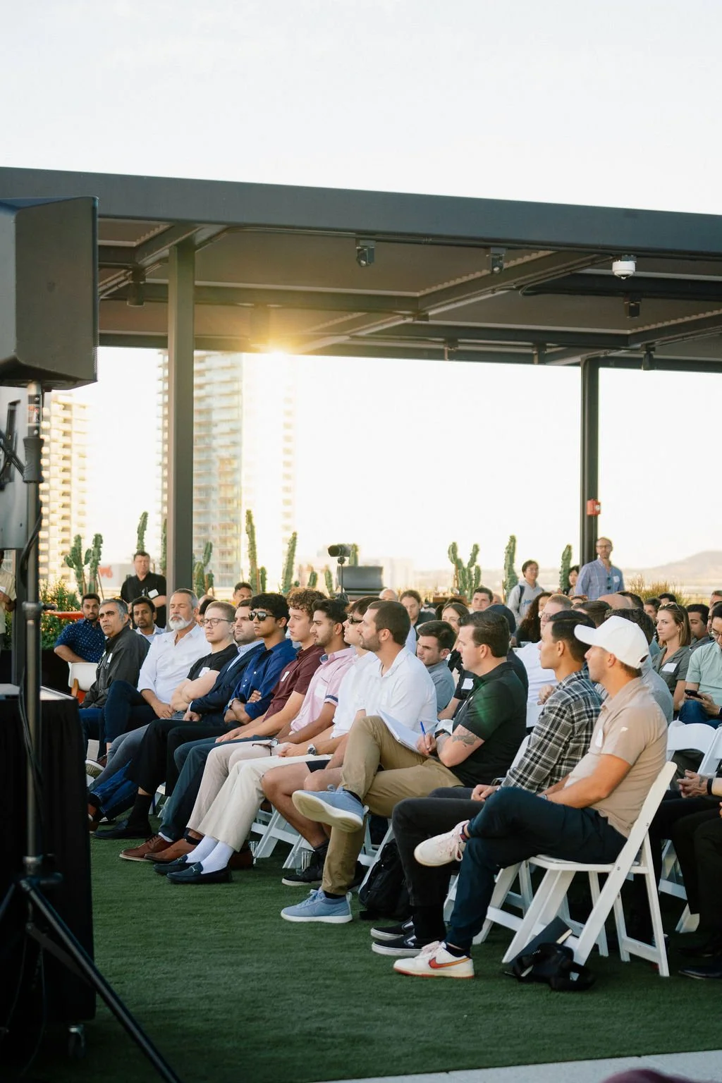An audience seated outdoors during a conference or event, with some taking notes, and tall buildings in the background at sunset.