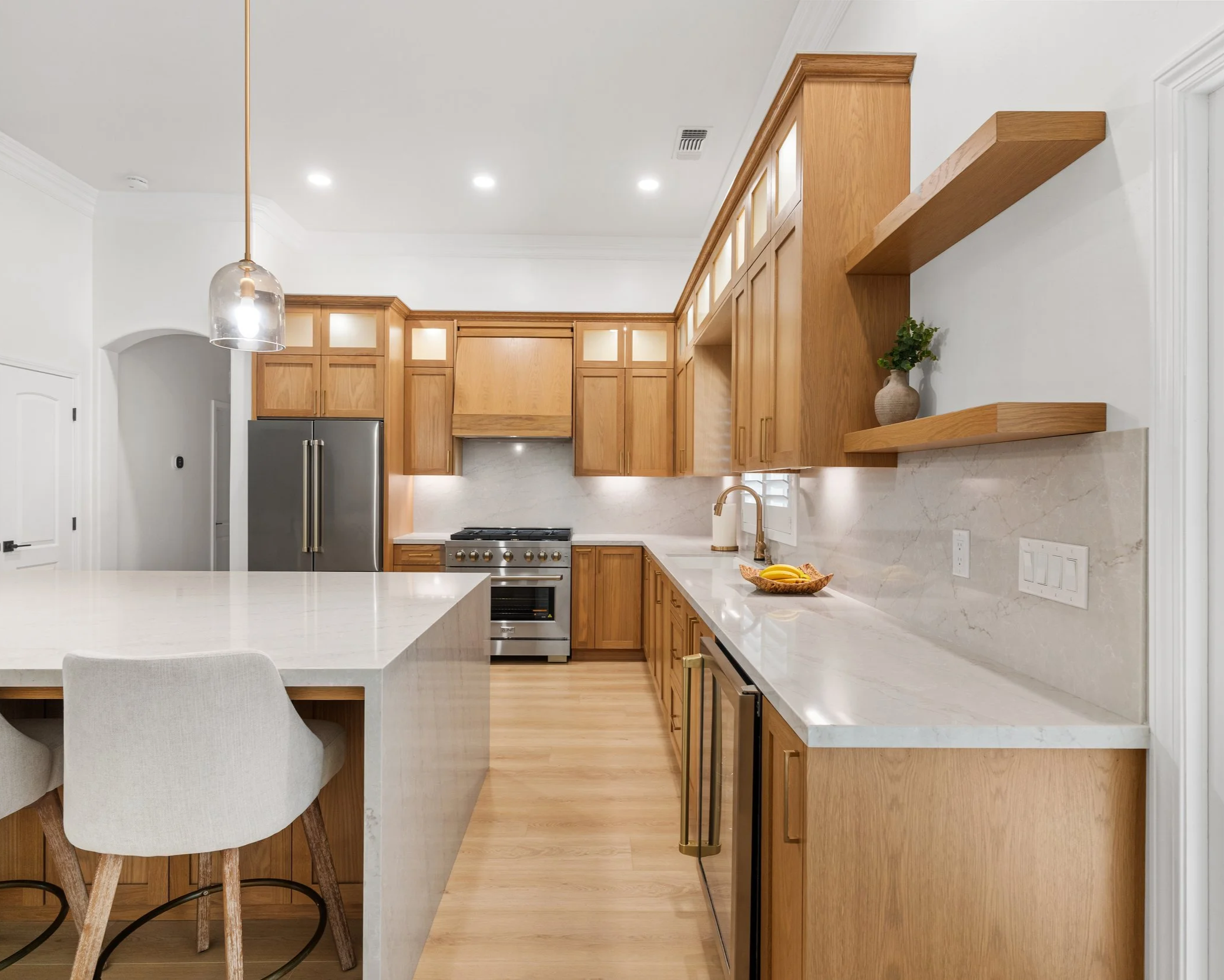 Modern kitchen with white marble countertops, wooden cabinets, a stainless steel refrigerator, and a large island. There is a pendant light hanging from the ceiling and a decorative plant in a vase on an open wooden shelf.