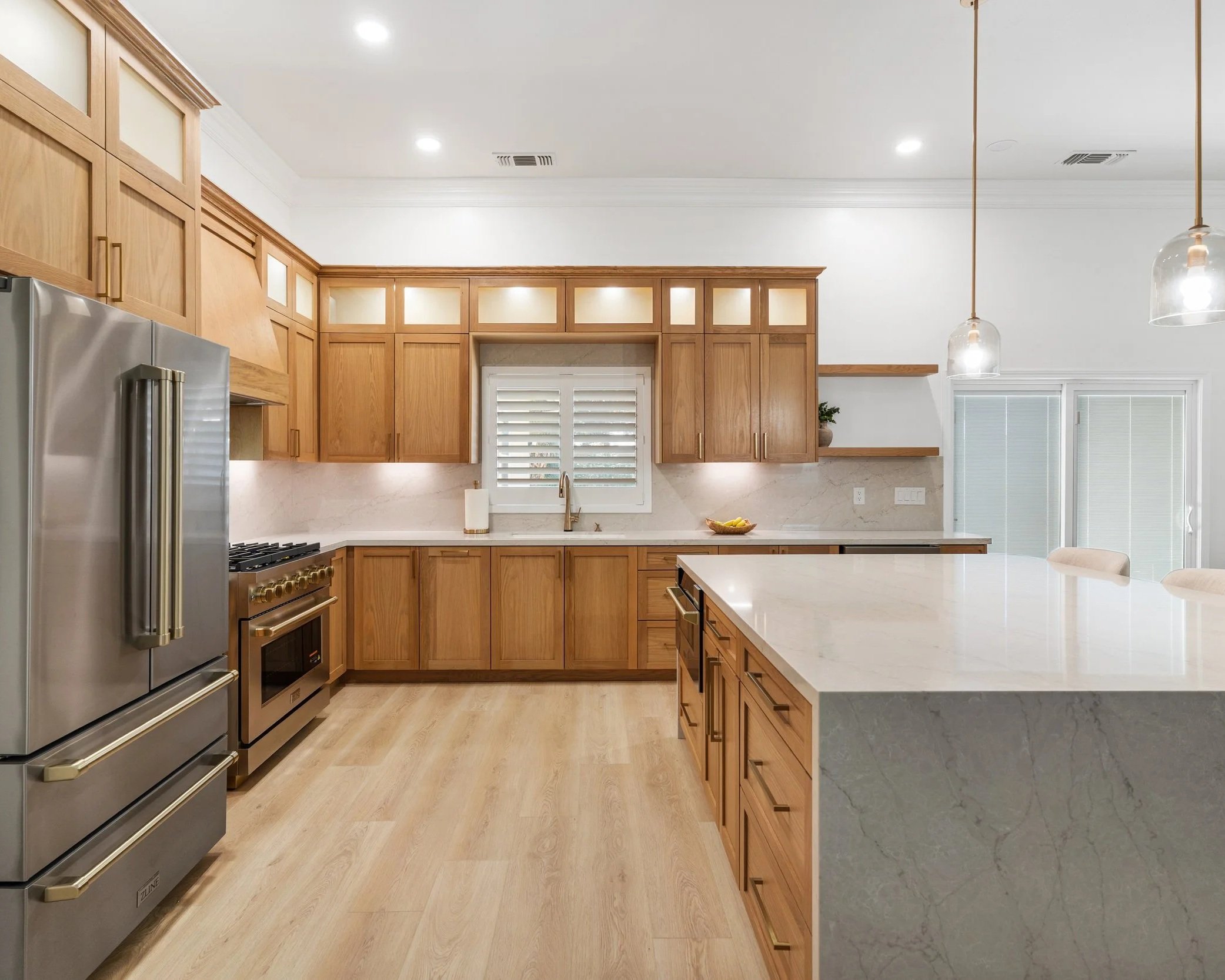 Modern kitchen with wooden cabinetry, stainless steel appliances, marble countertops, pendant lights, and natural light from window with shutters.