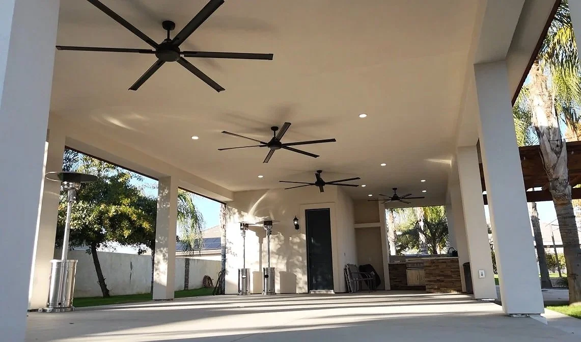 Covered outdoor patio with ceiling fans, recessed lighting, and outdoor heaters, surrounded by trees and a white fence.