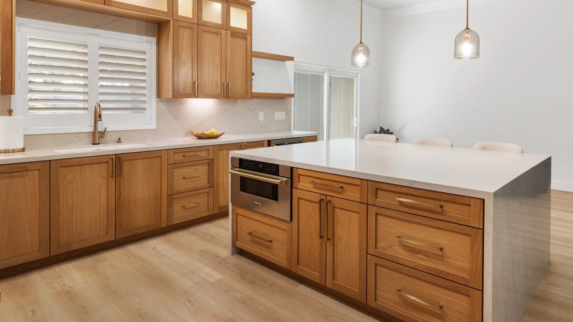 Modern kitchen with wooden cabinets, marble countertops, a large central island, stainless steel oven, black dishwasher, window with shutters, pendant lights, and light wood flooring.
