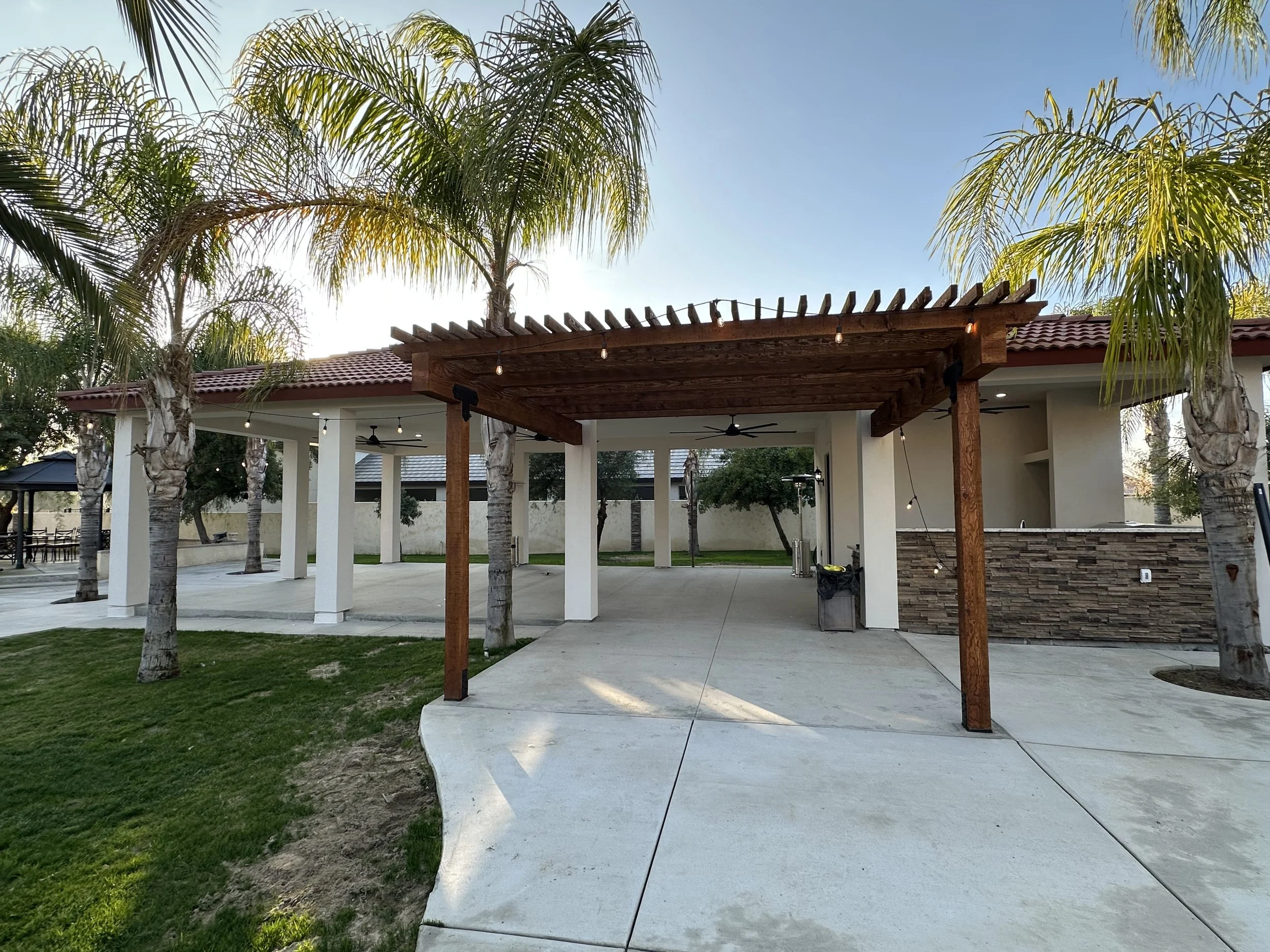A backyard patio area with a pergola, palm trees, string lights, ceiling fans, and a grass lawn.