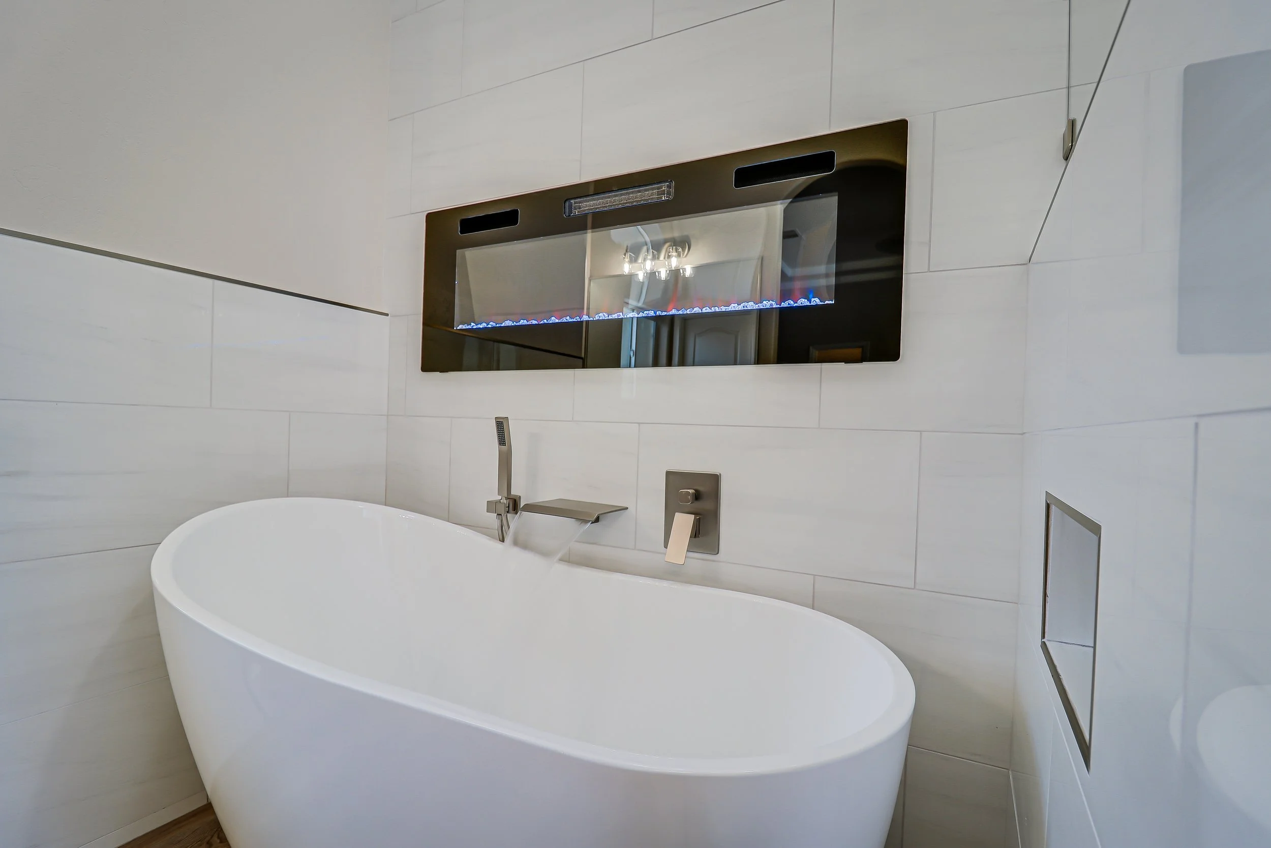 A modern bathroom with a white freestanding bathtub, a wall-mounted faucet, a large mirror, and a wall with light-colored tiles.