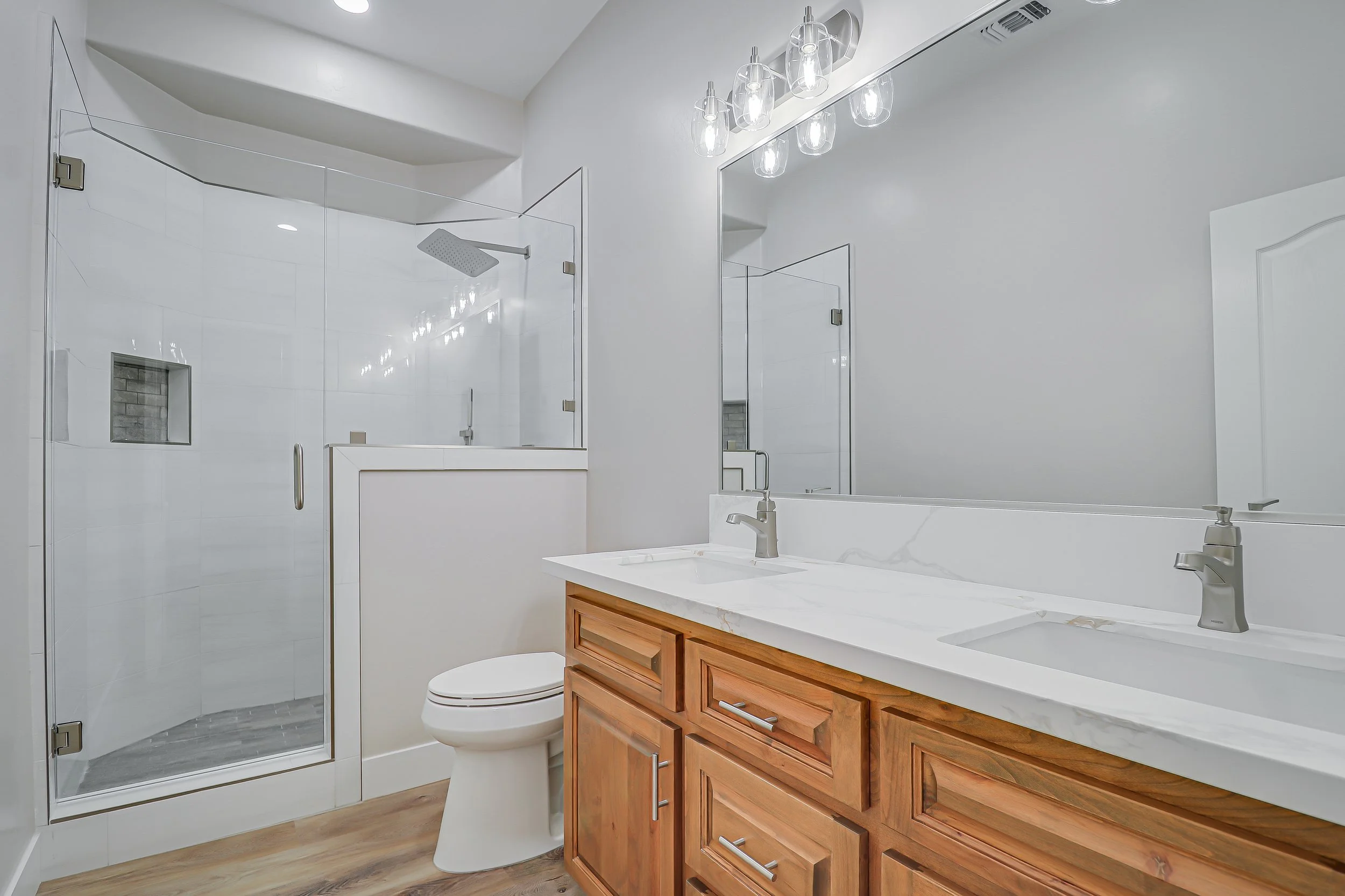 Modern bathroom with a walk-in shower, white walls, a wooden vanity with double sinks, a large mirror, and a ceiling light fixture.