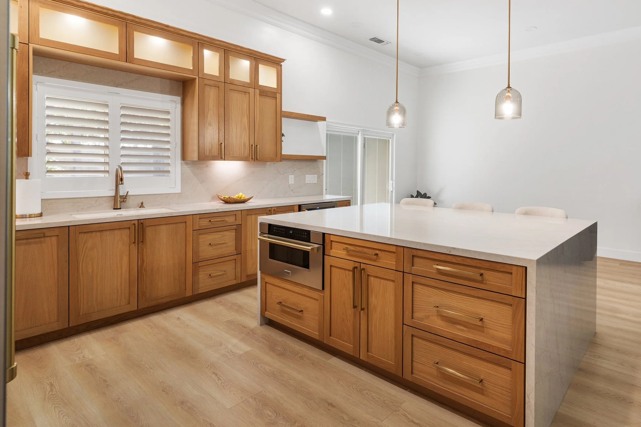 A modern kitchen with wooden cabinets, a white marble countertop, and stainless steel appliances. Two pendant lights hang above the island, with a window and sliding door in the background.