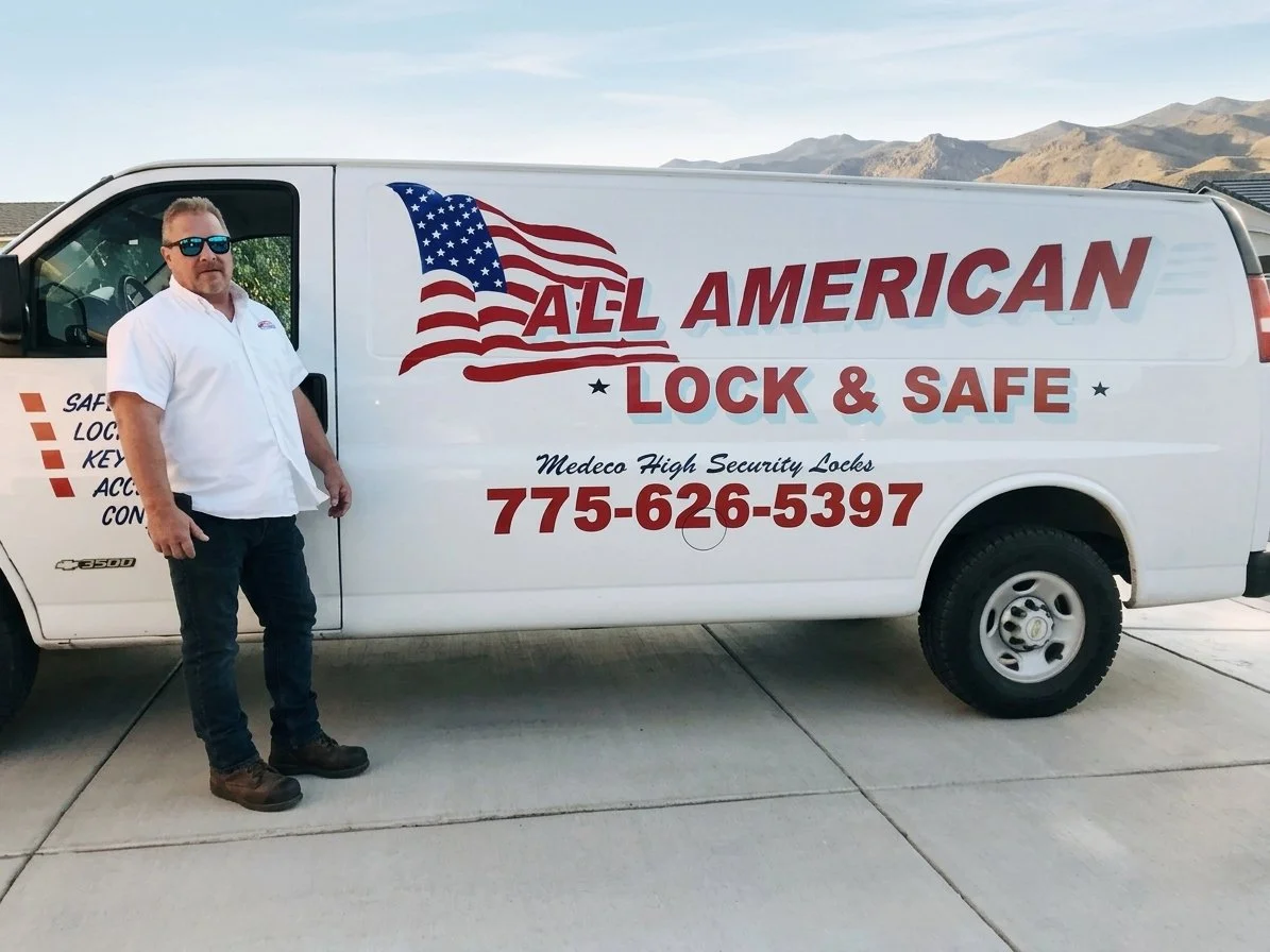 A man wearing sunglasses and a white shirt standing next to a white work van with red and blue lettering and an American flag graphic promoting American Lock & Safe, a security locksmith company. The van displays contact number 775-626-5397 and the text 'Medeco High Security Locks'. Mountains are visible in the background.