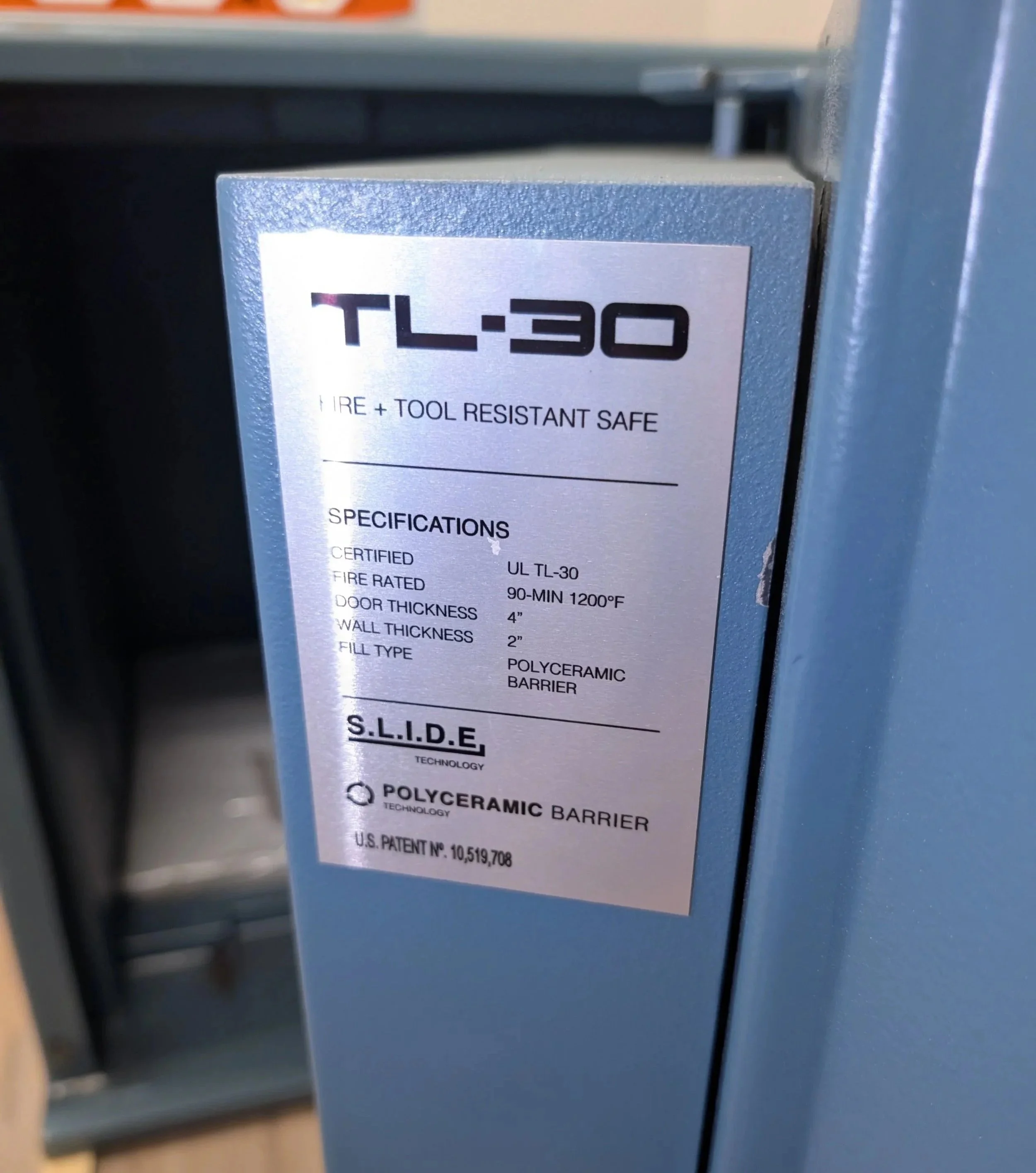 Close-up of a silver fire resistant safe with a label showing specifications, mounted on a blue wall or shelf in an industrial or storage setting.