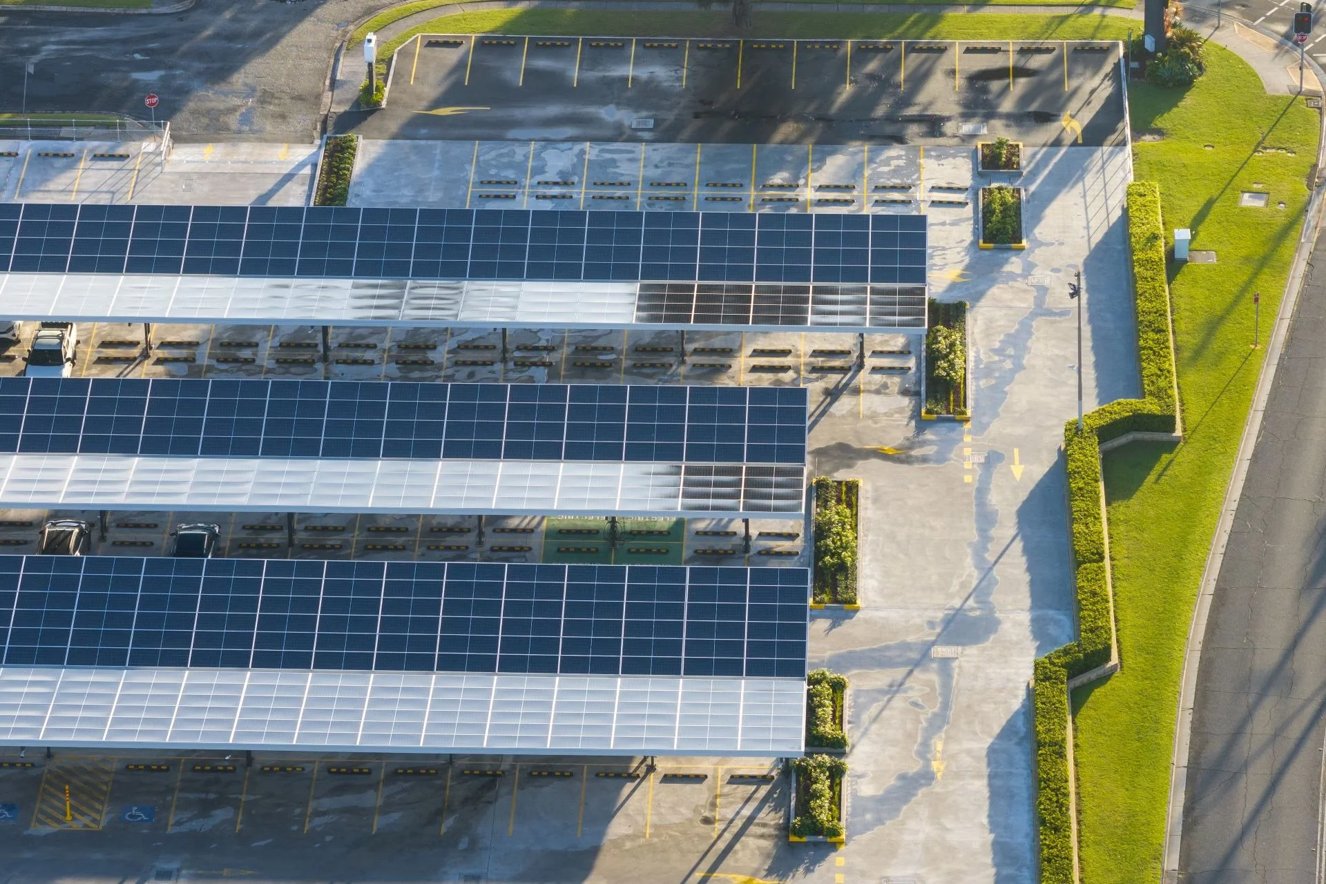 An aerial view of a parking lot with solar panel-covered carports, surrounded by green grass, small trees, and sidewalks.
