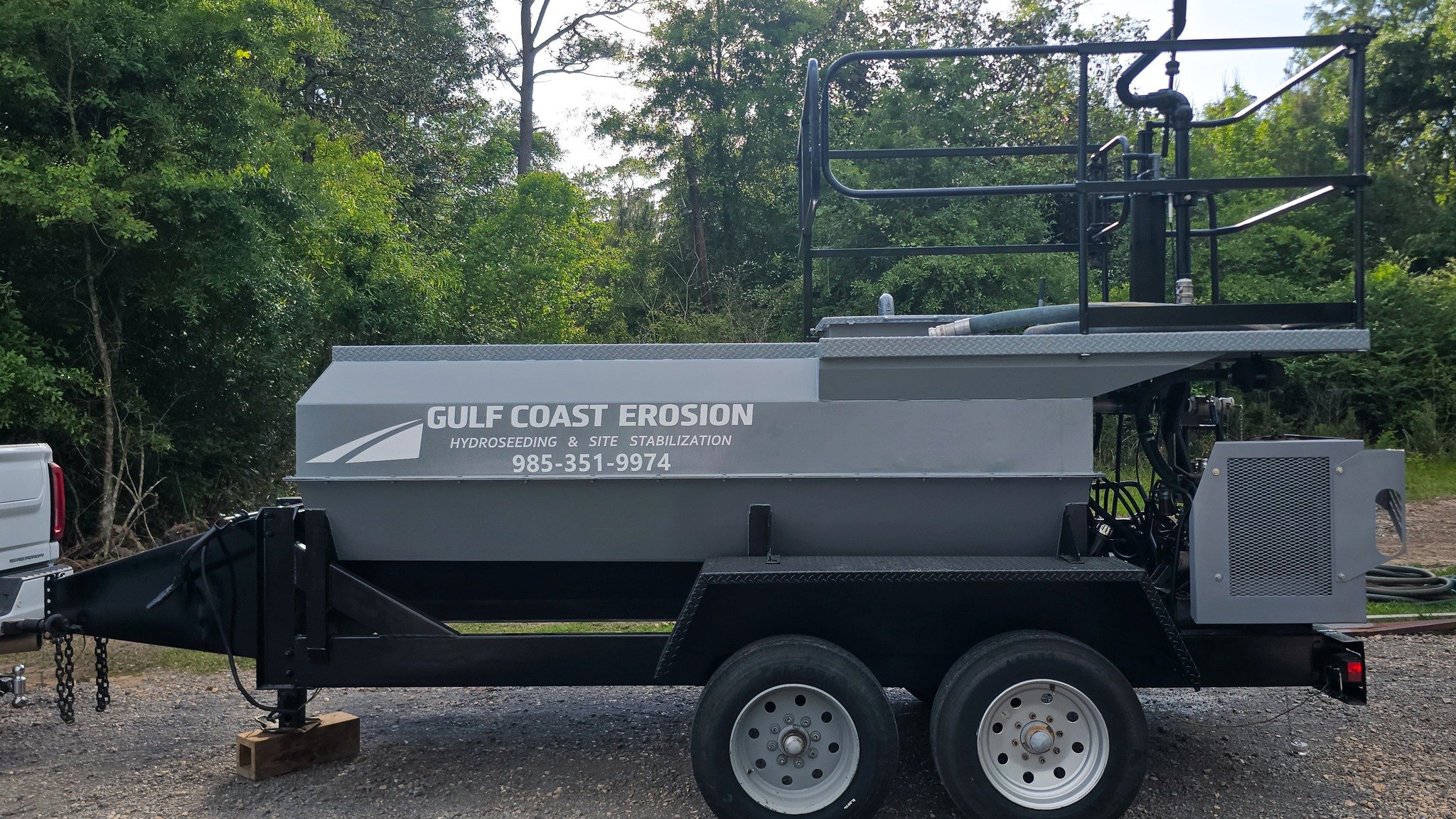 A trailer with the label 'Gulf Coast Erosion Hydroseeding & Site Stabilization' attached to a truck, parked on a gravel area with green trees in the background.