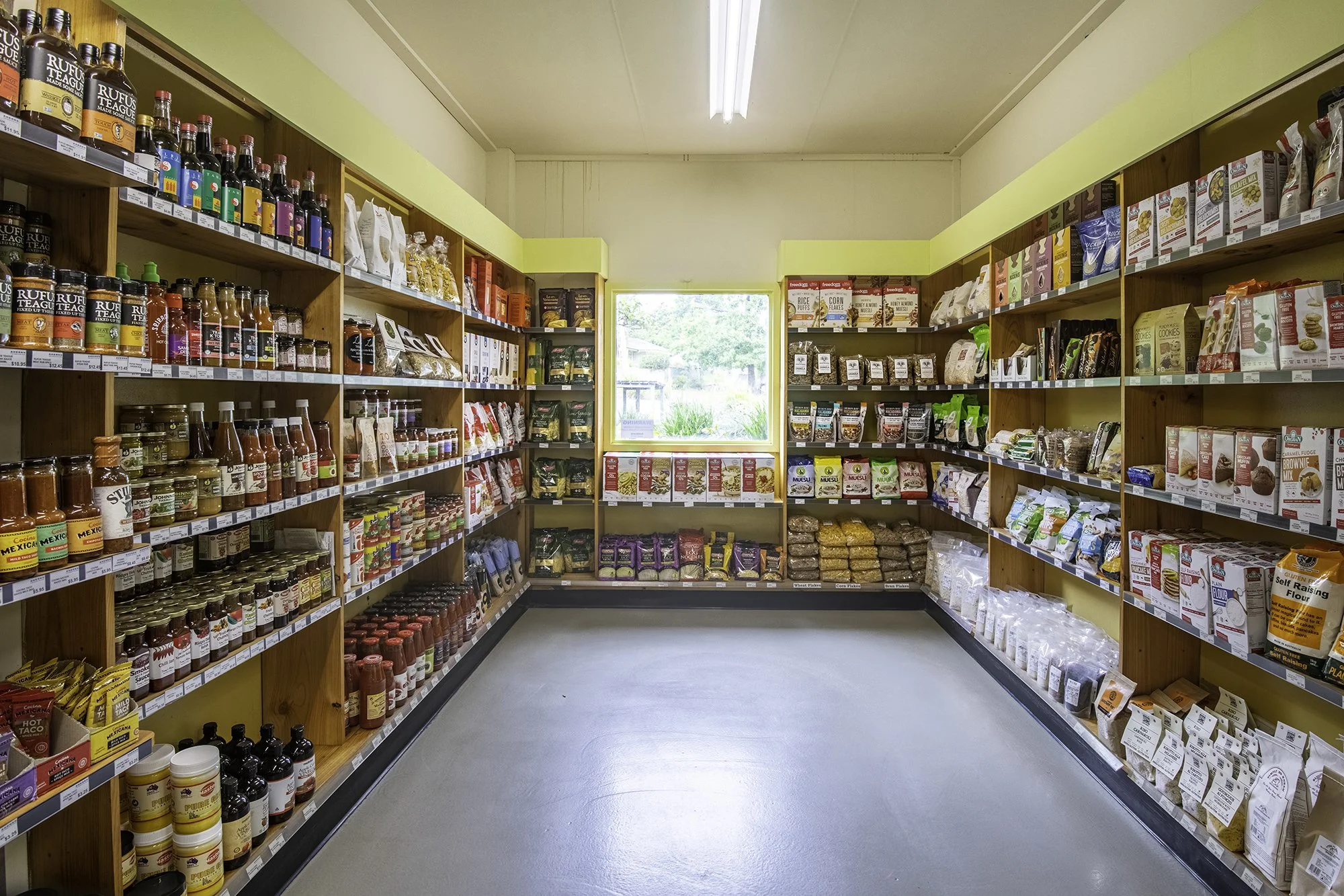 Inside a grocery store aisle with wooden shelves filled with various food products such as sauces, grains, and baking ingredients, with a window at the end showing outdoor greenery.