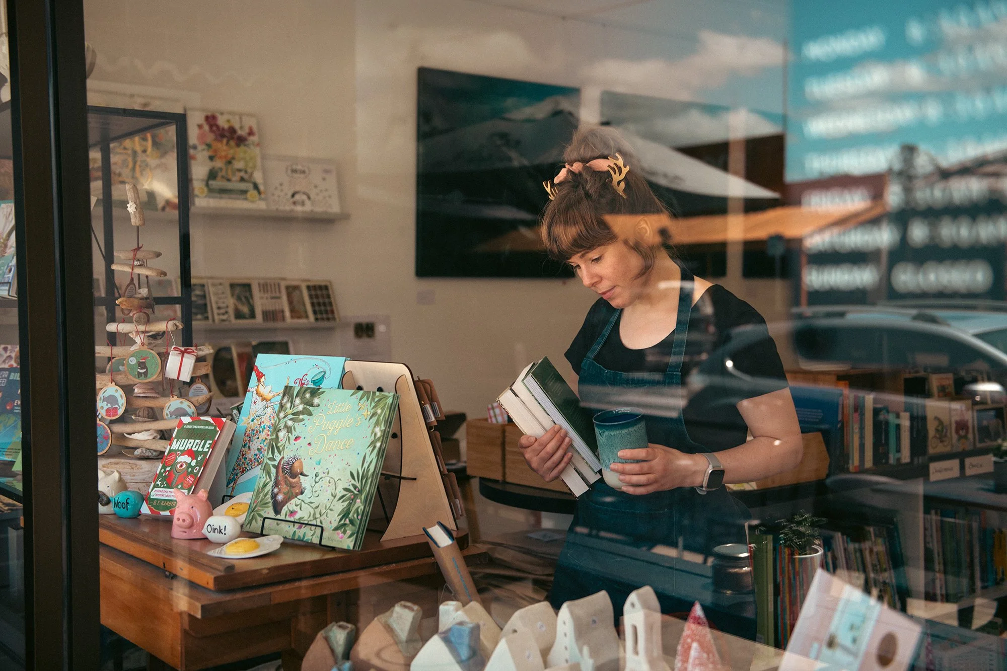 Woman shopping for books inside a bookstore, viewed through a glass window. She is holding a book and a cup, wearing a black top and a watch, with decorative hairpins. The display table has children's books and small toys, with the book titles 'Littl