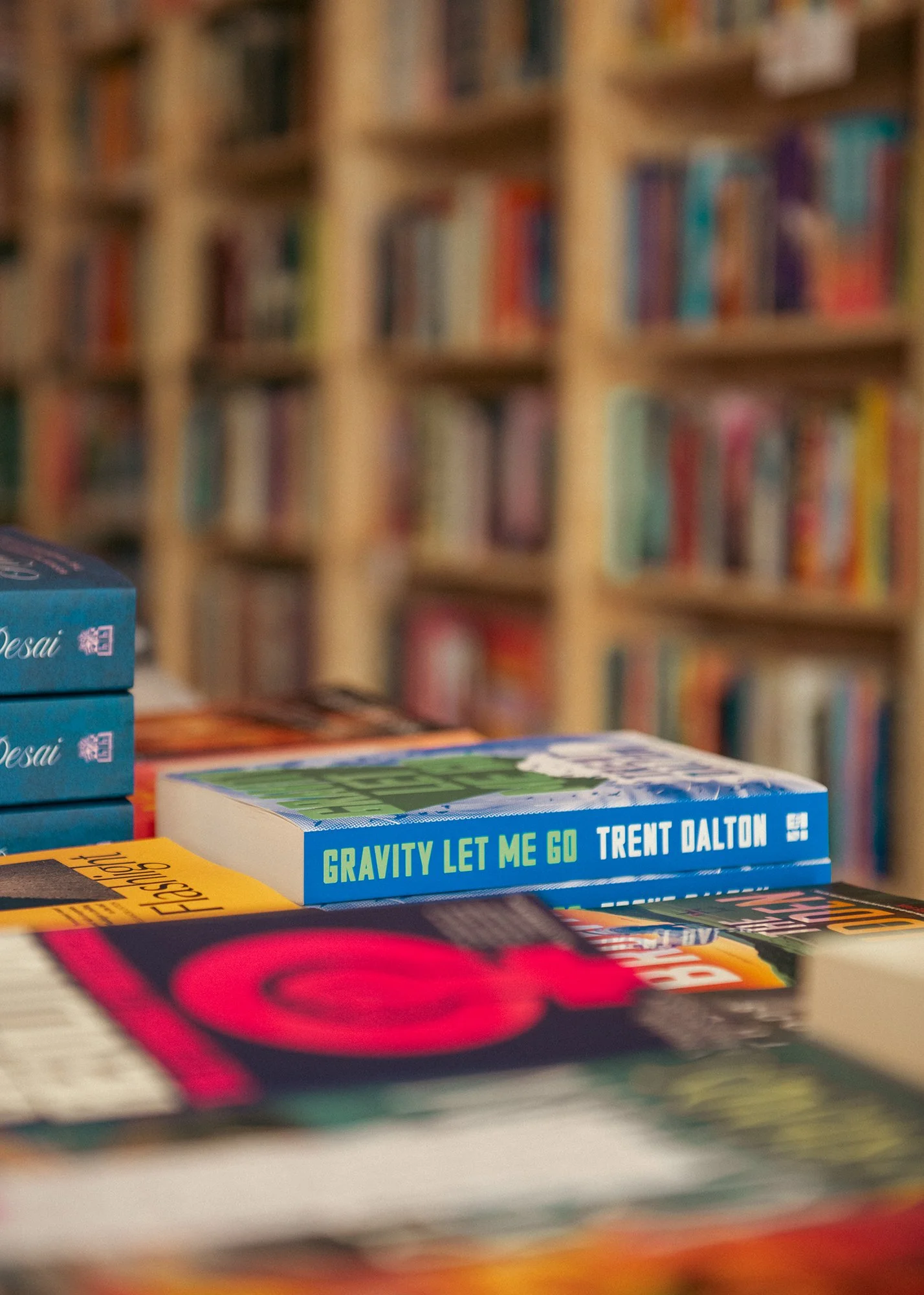 Blurred bookshelf in the background with a close-up of a book titled 'Gravity Let Me Go' by Trent Dalton in the foreground.