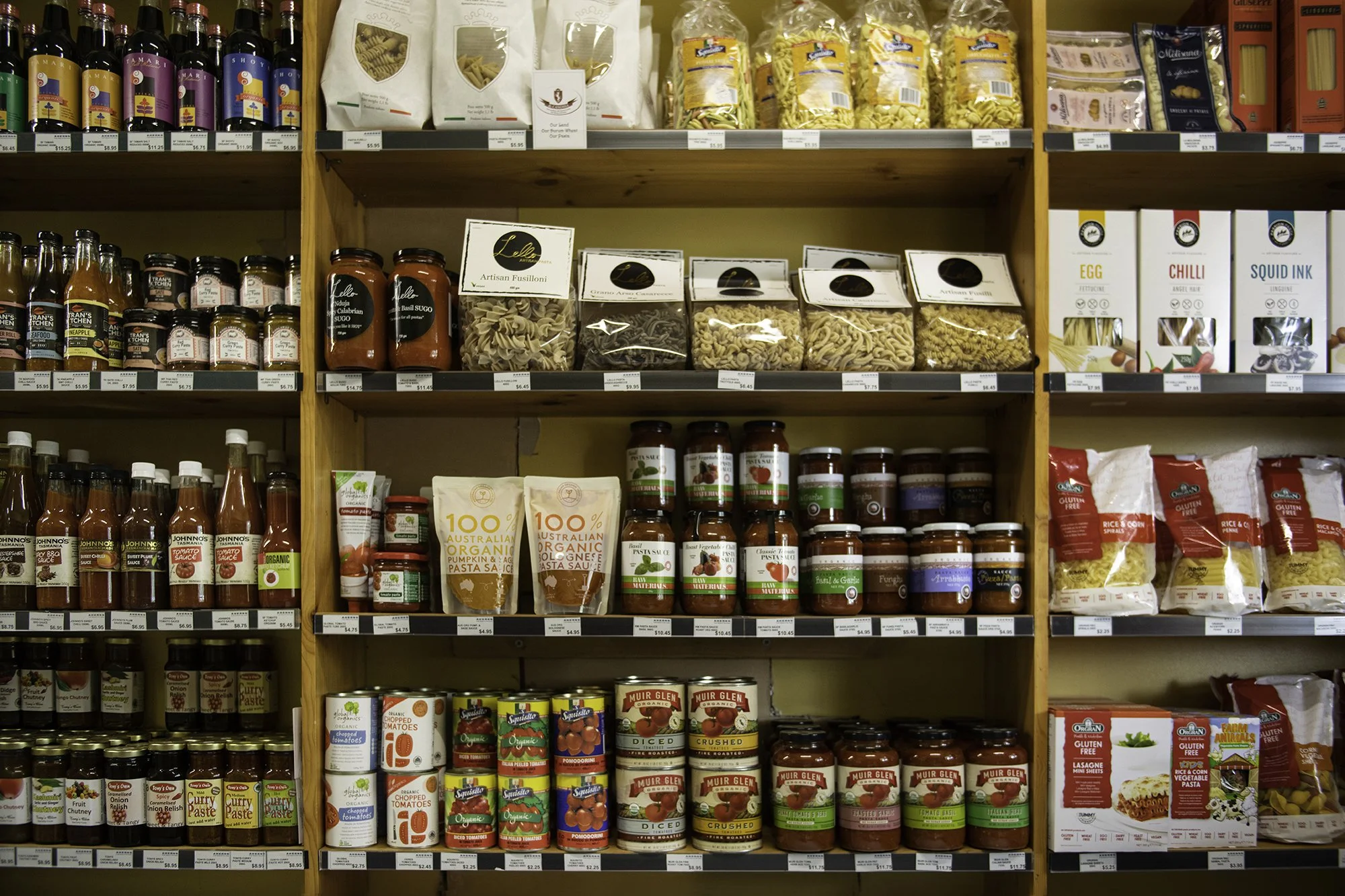 Shelf in a grocery store with various pasta, pasta sauces, and other packaged food items. Top shelf has wine bottles and pasta packets. Middle shelves contain jars of sauces and canned vegetables. Bottom shelf features canned diced, crushed, and chop