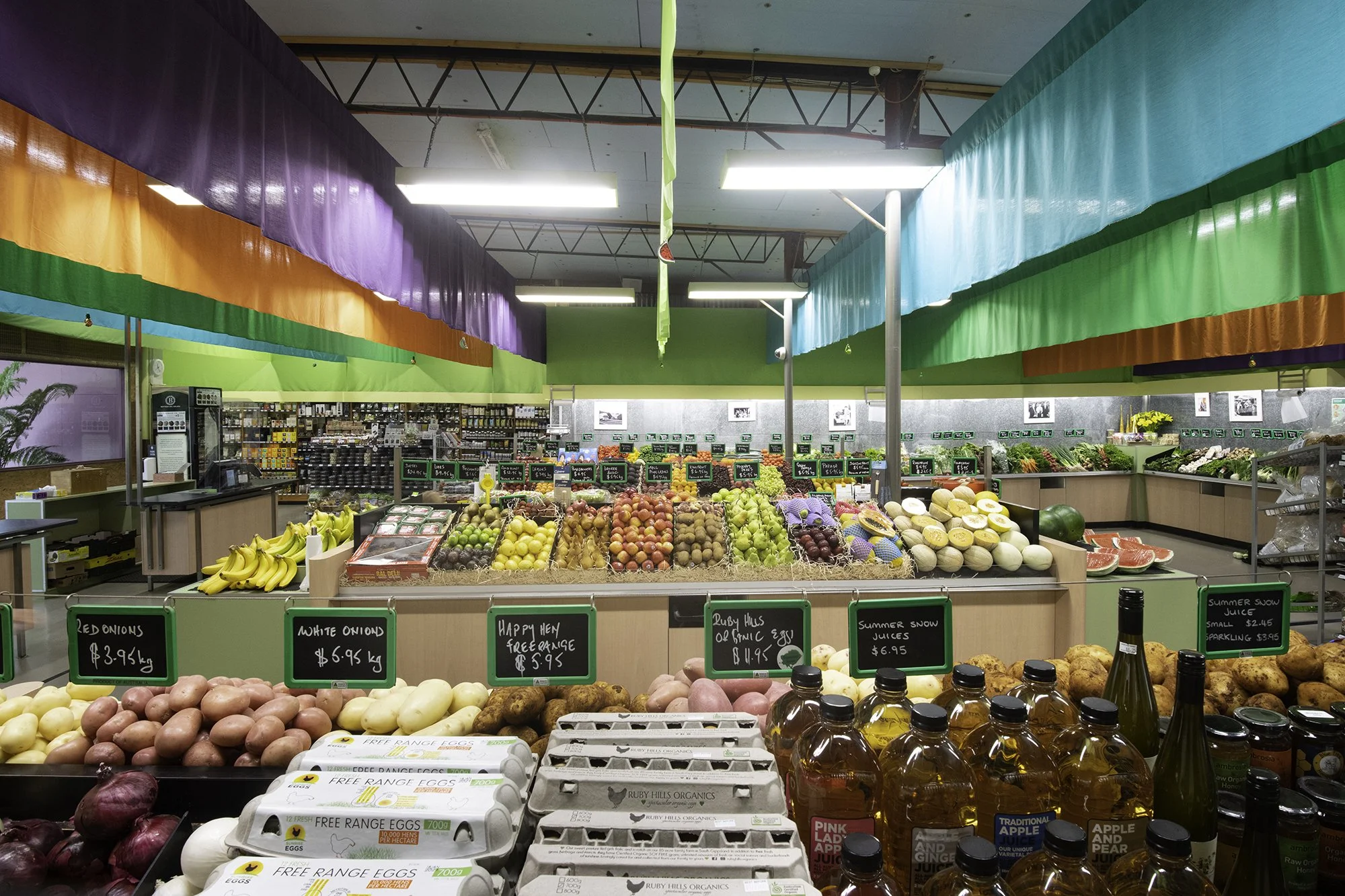 A grocery store produce section with various fruits and vegetables on display, including onions, eggs, and bottles of juice, under colorful fabric decorations on the ceiling.
