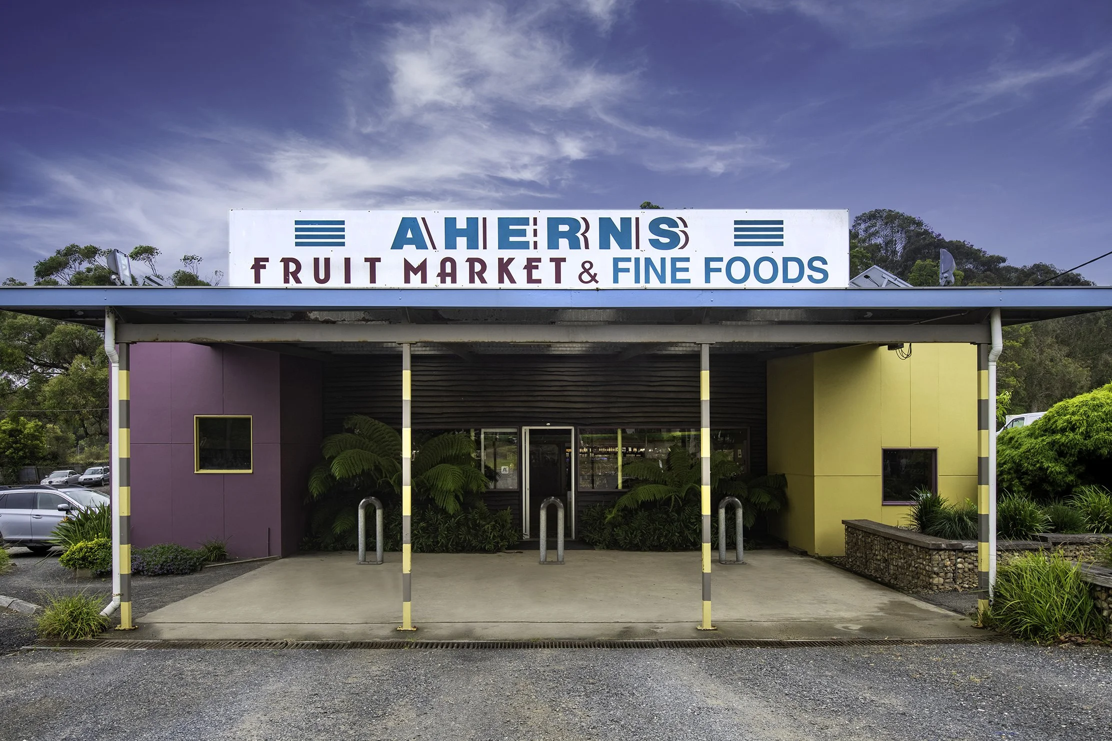 Sign for Aherns Fruit Market and Fine Foods on top of a building with colorful exterior walls, surrounded by greenery, parking lot, and a blue sky with some clouds.