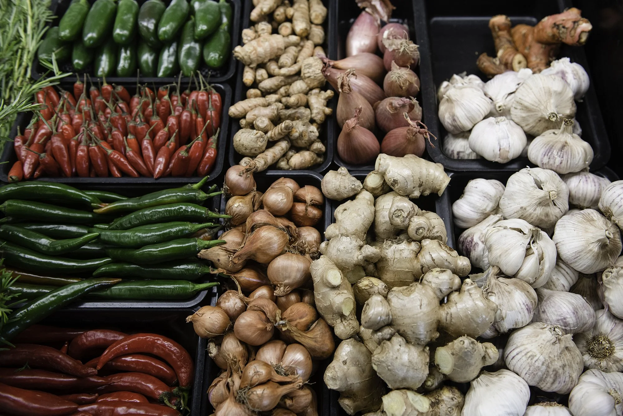 A display of fresh vegetables including red chili peppers, green chili peppers, ginger, shallots, garlic, and turmeric arranged in black trays at a market.