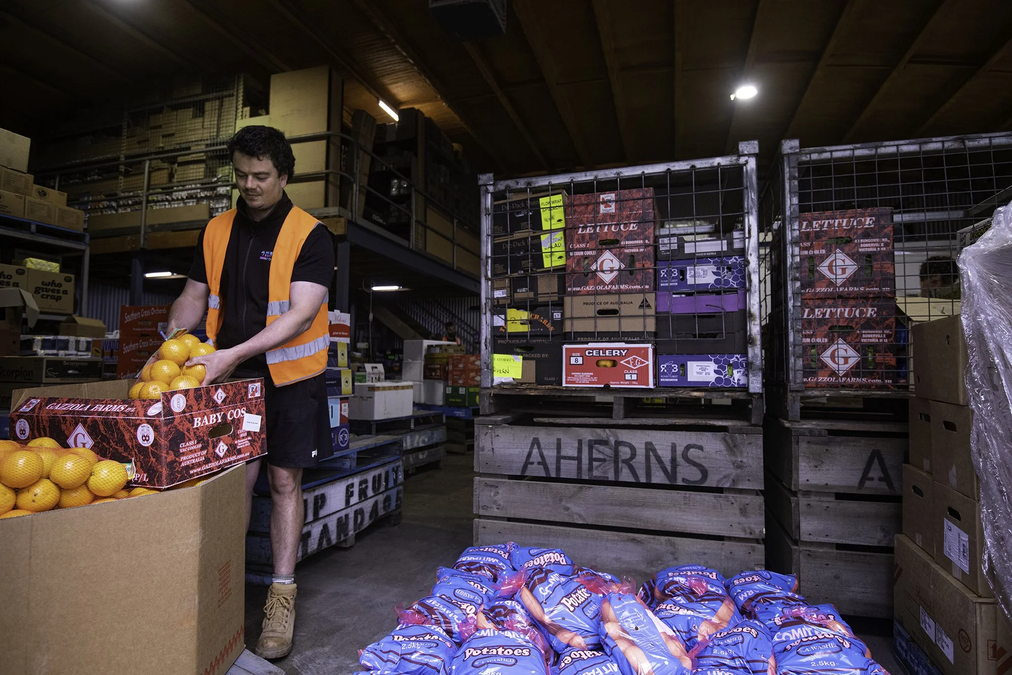 A man in an orange safety vest and black shirt is stacking oranges in a cardboard box inside a warehouse or grocery store. Storage shelves with boxes and bags of produce are visible in the background.