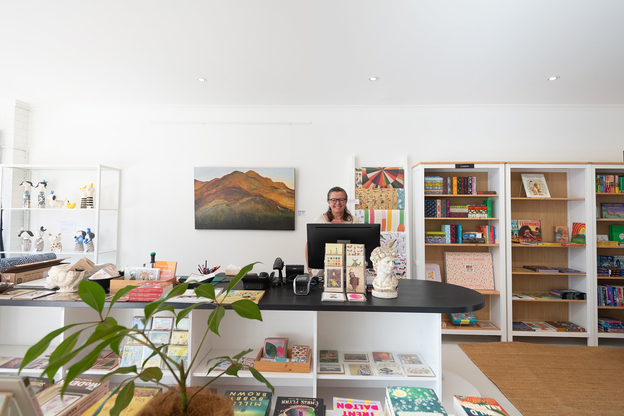 Interior of a bookstore or library with white walls and bookshelves filled with books. A woman with glasses and long hair is standing behind a cashier counter with a black top, smiling at the camera. There are art pieces, a sculpture, and various ite