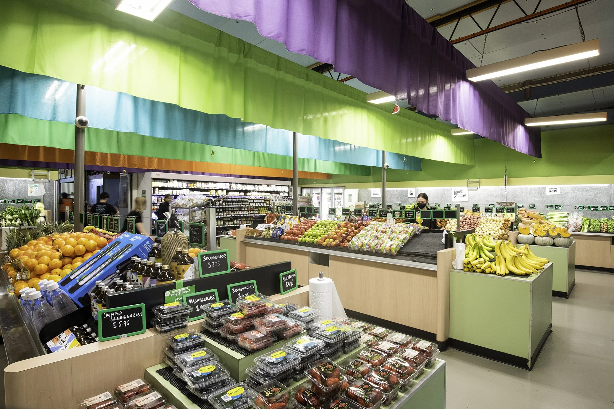 A grocery store produce section with colorful fabric drapes hanging from the ceiling, displaying fruits such as oranges, strawberries, blueberries, bananas, and other produce on wooden displays, and a few customers shopping.