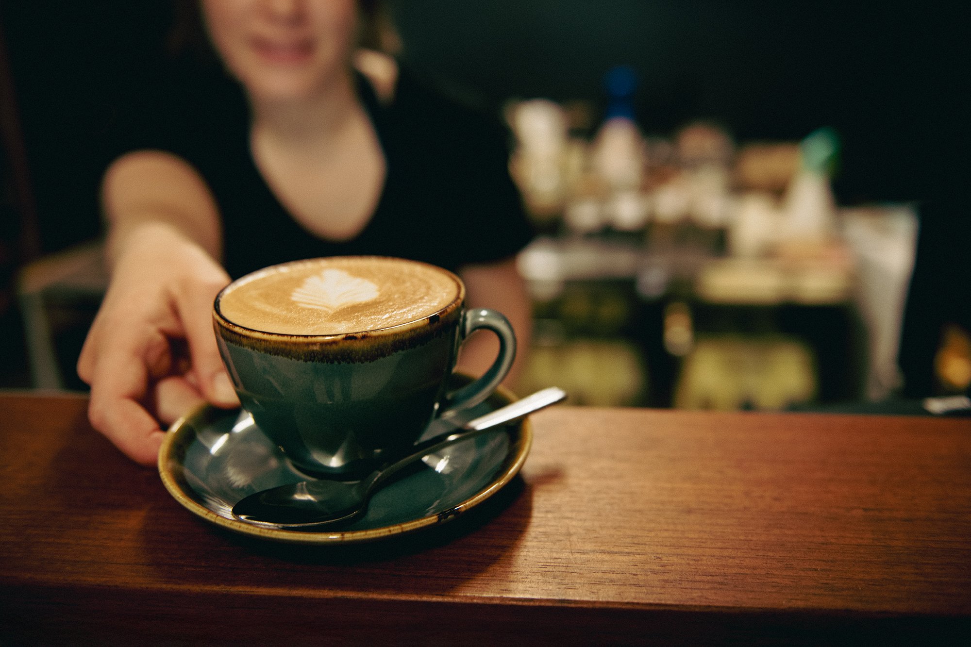 A person holds a dark ceramic cup of latte with latte art, sitting on a matching saucer with a spoon, at a wooden table in a cafe. In the background, there are several bottles and containers, slightly blurred.
