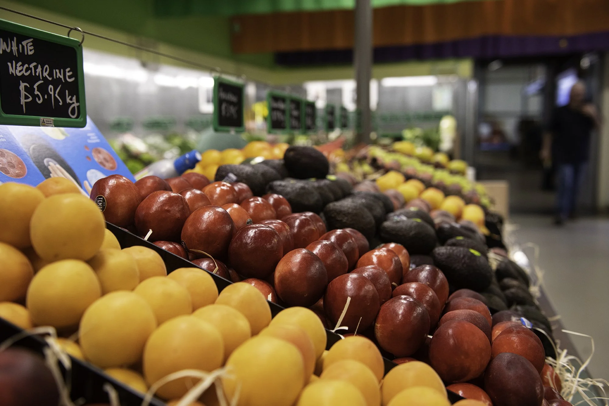 Fresh fruits on display at a grocery store, including oranges, red apples, and avocados, with a person browsing in the background.