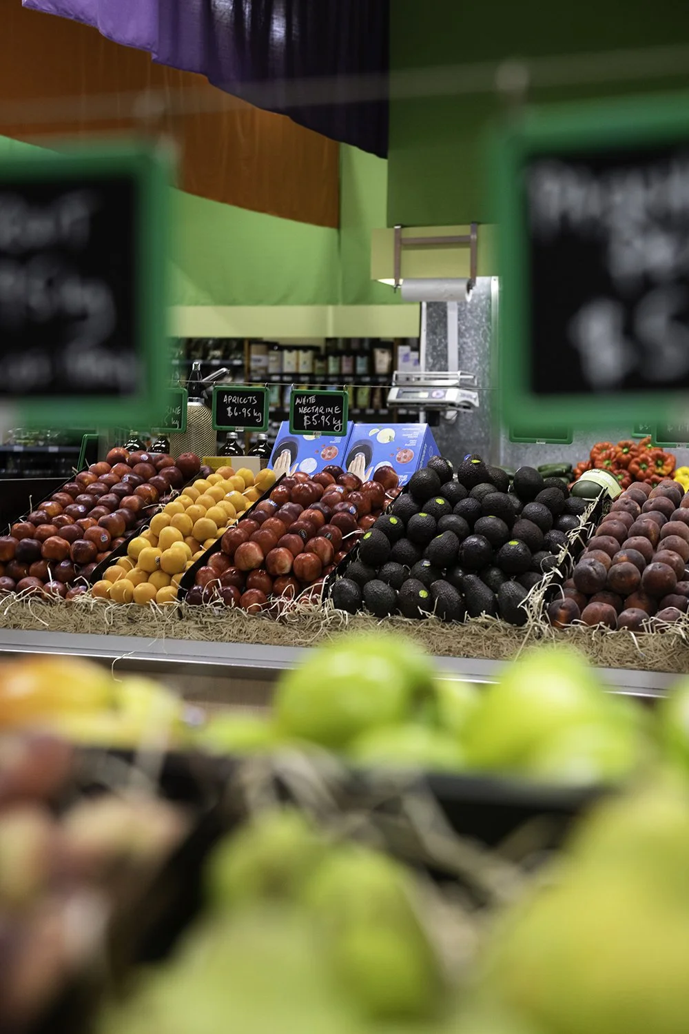 A display of fresh fruits including apricots, red apples, black avocados, and peaches at a grocery store.