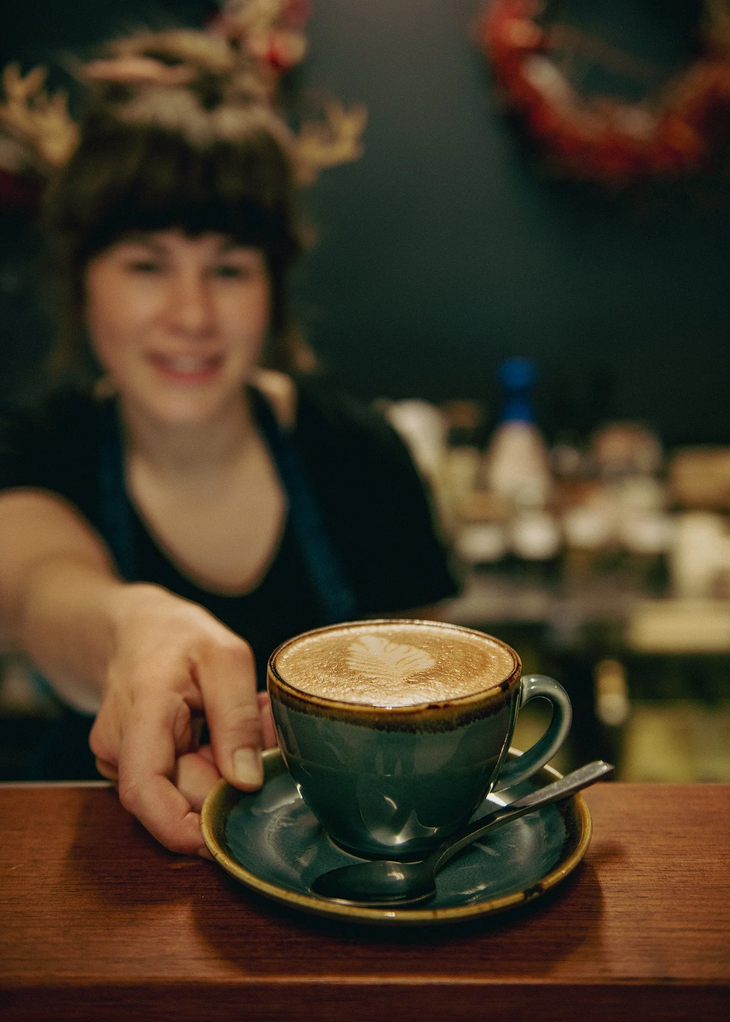 A person extending their arm to hand a cup of coffee to someone, with a smile and blurry face in the background. The coffee is in a green ceramic cup on a matching saucer, with a spoon resting on the saucer, placed on a wooden table.