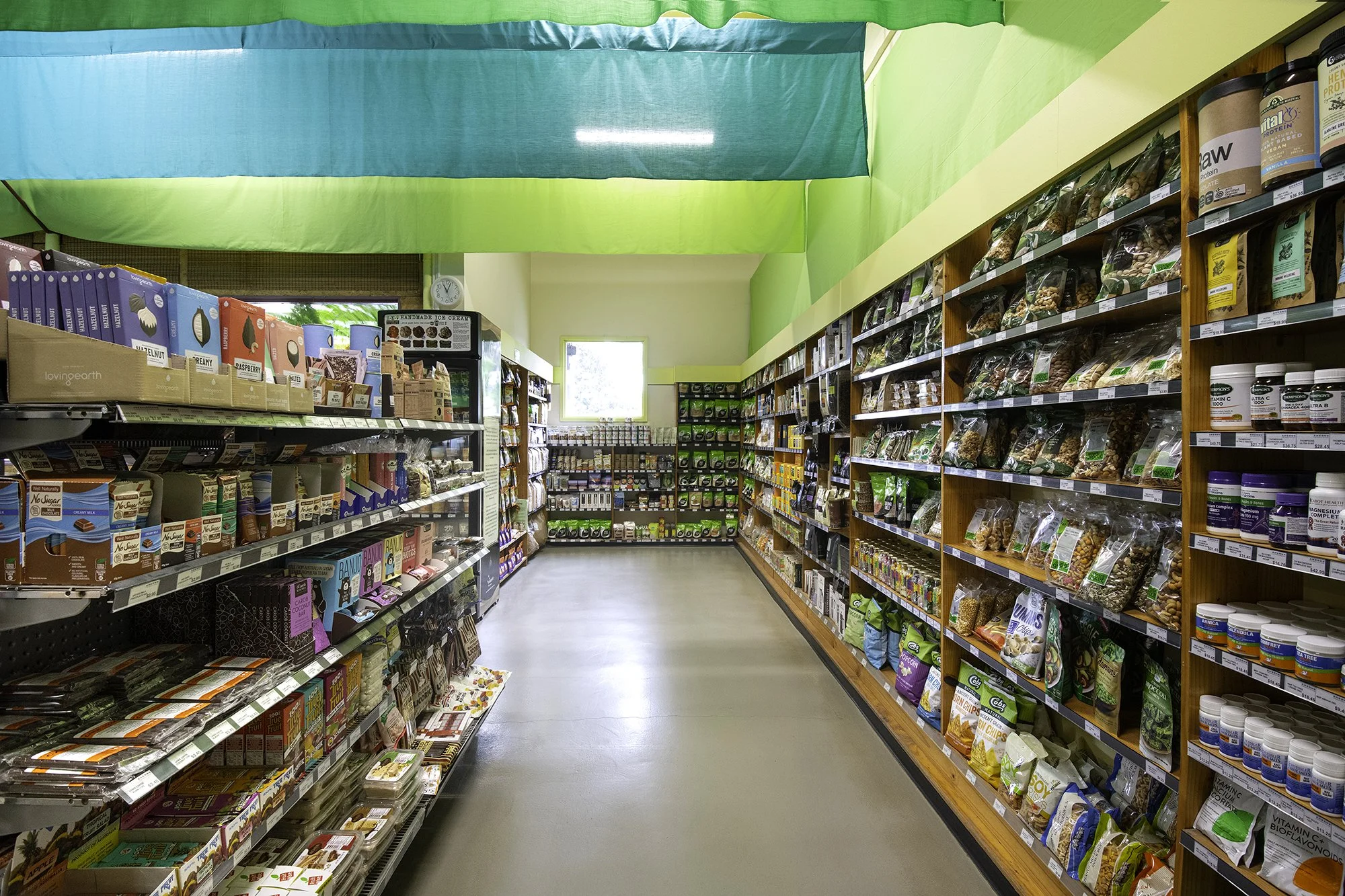 Inside a grocery store aisle with shelves stocked with snacks, supplements, and health products, colorful green and blue fabric ceiling decorations, a small window, and a clock on the wall.
