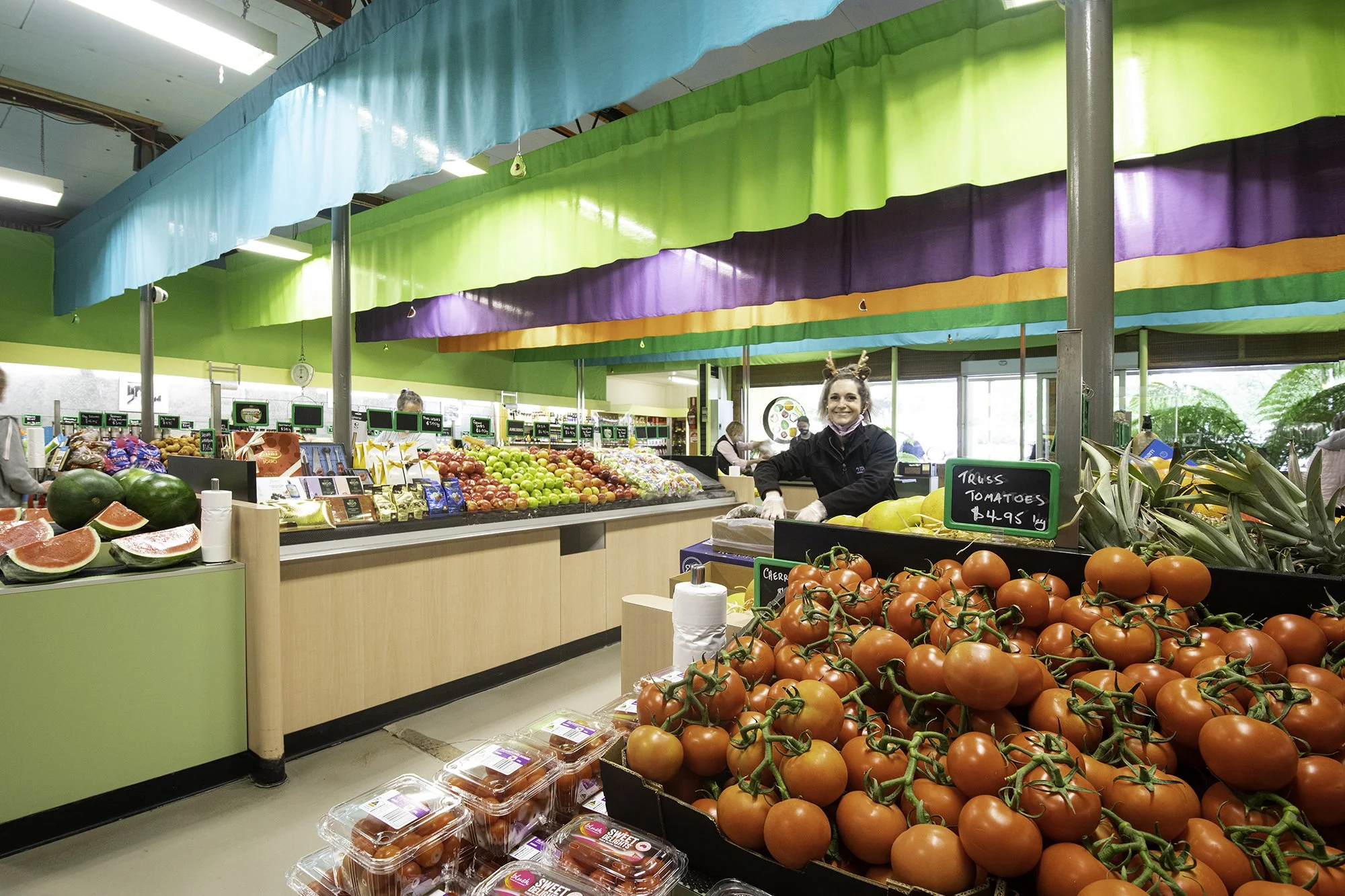 Inside a grocery store with fresh produce displays, including tomatoes, watermelons, green apples, and packaged strawberries. Colorful fabric decorations hang from the ceiling, and store employees are visible in the background.