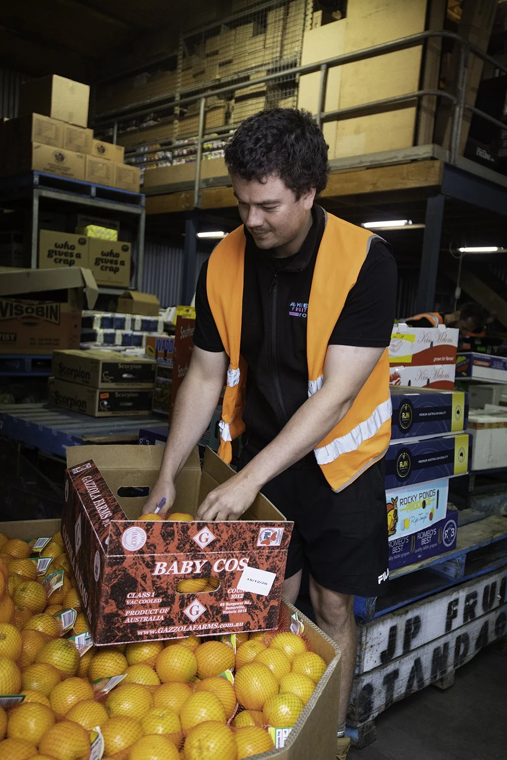 A man in a black shirt and orange safety vest packing oranges into a cardboard box in a warehouse or produce storage area.