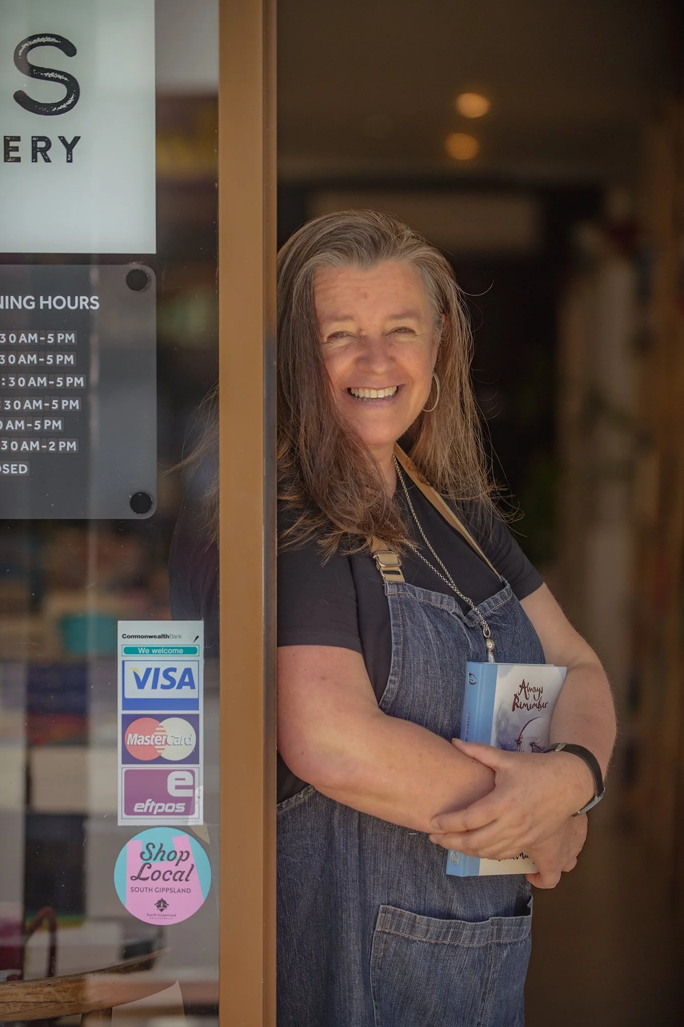 A woman smiling and standing inside a store, holding a book and wearing a denim apron, with a glass door and store signs visible.