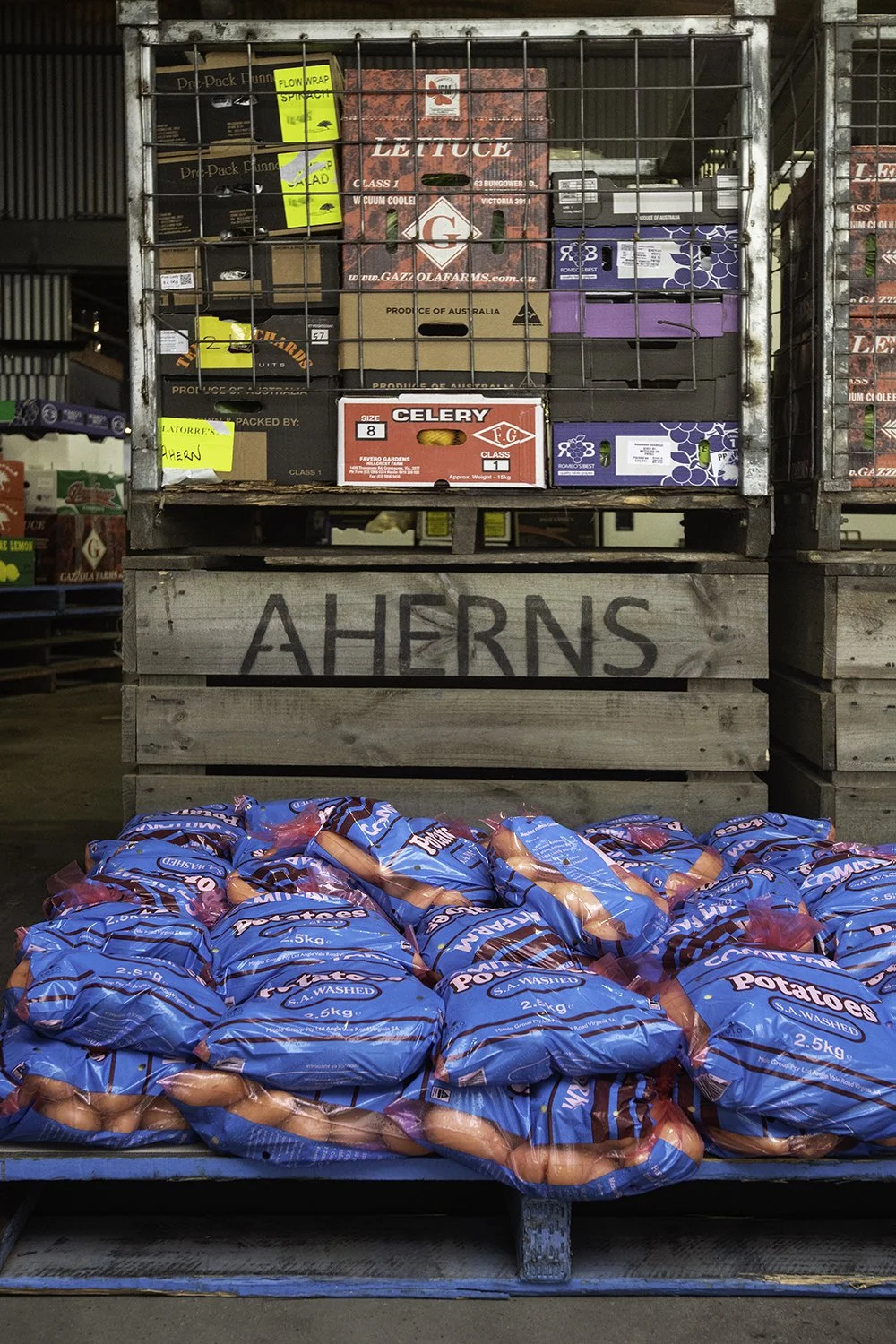 A display of potatoes in blue bags on a blue shelf at a grocery store, with crates of assorted produce and boxes of fruit stacked behind wooden pallets labeled "Aherns."