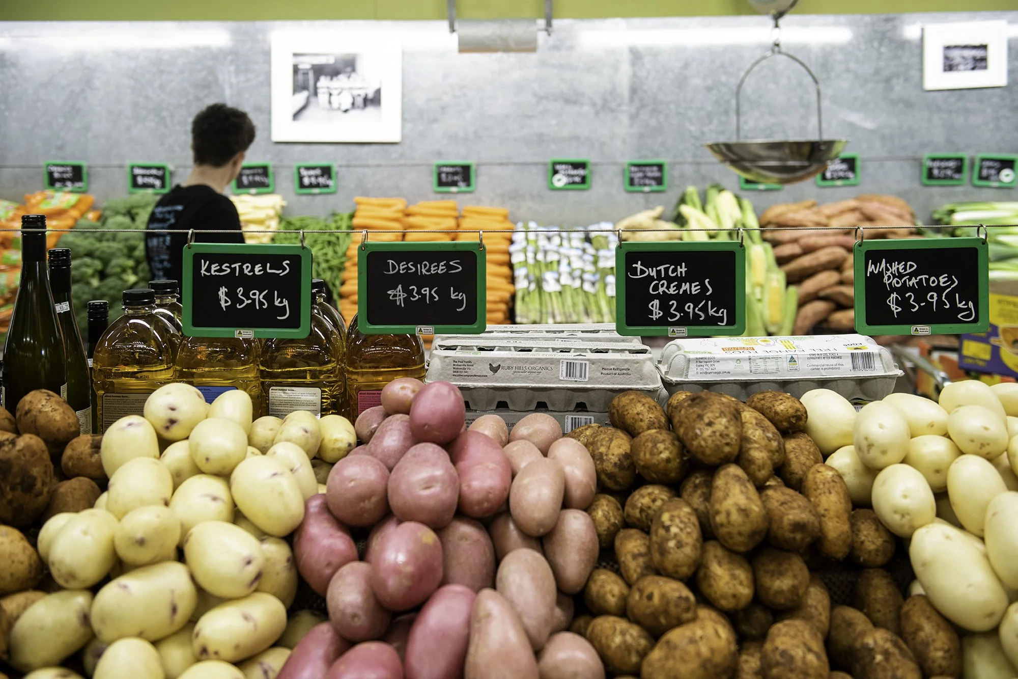 View of fresh produce at a grocery store, including apples, sweet potatoes, and agricultural bottles. In the background, there are carrots, celery, and sweet potatoes, with blackboard signs listing prices.