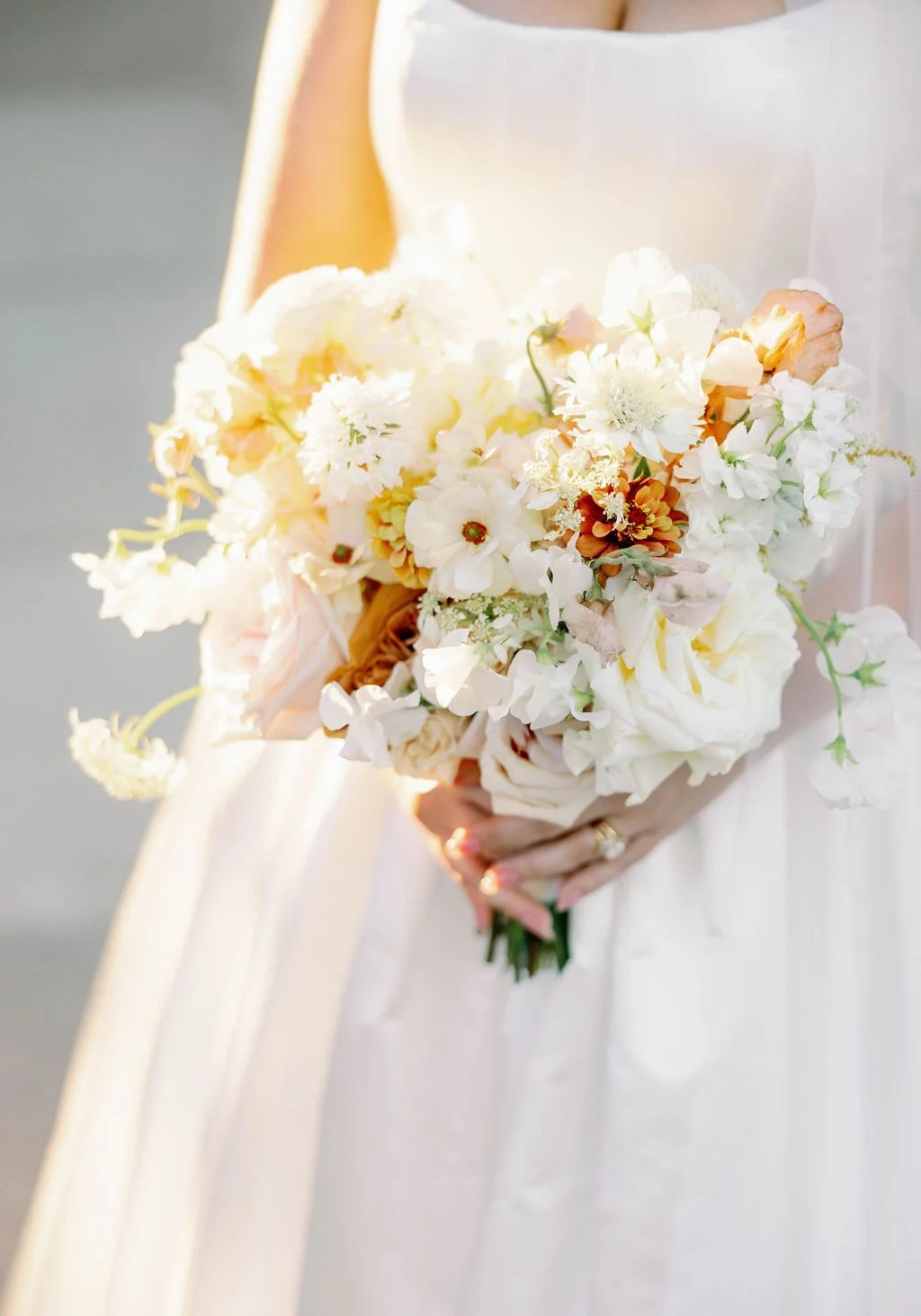 A bride holding a bouquet of white and light peach flowers, with some green foliage, against a soft background.
