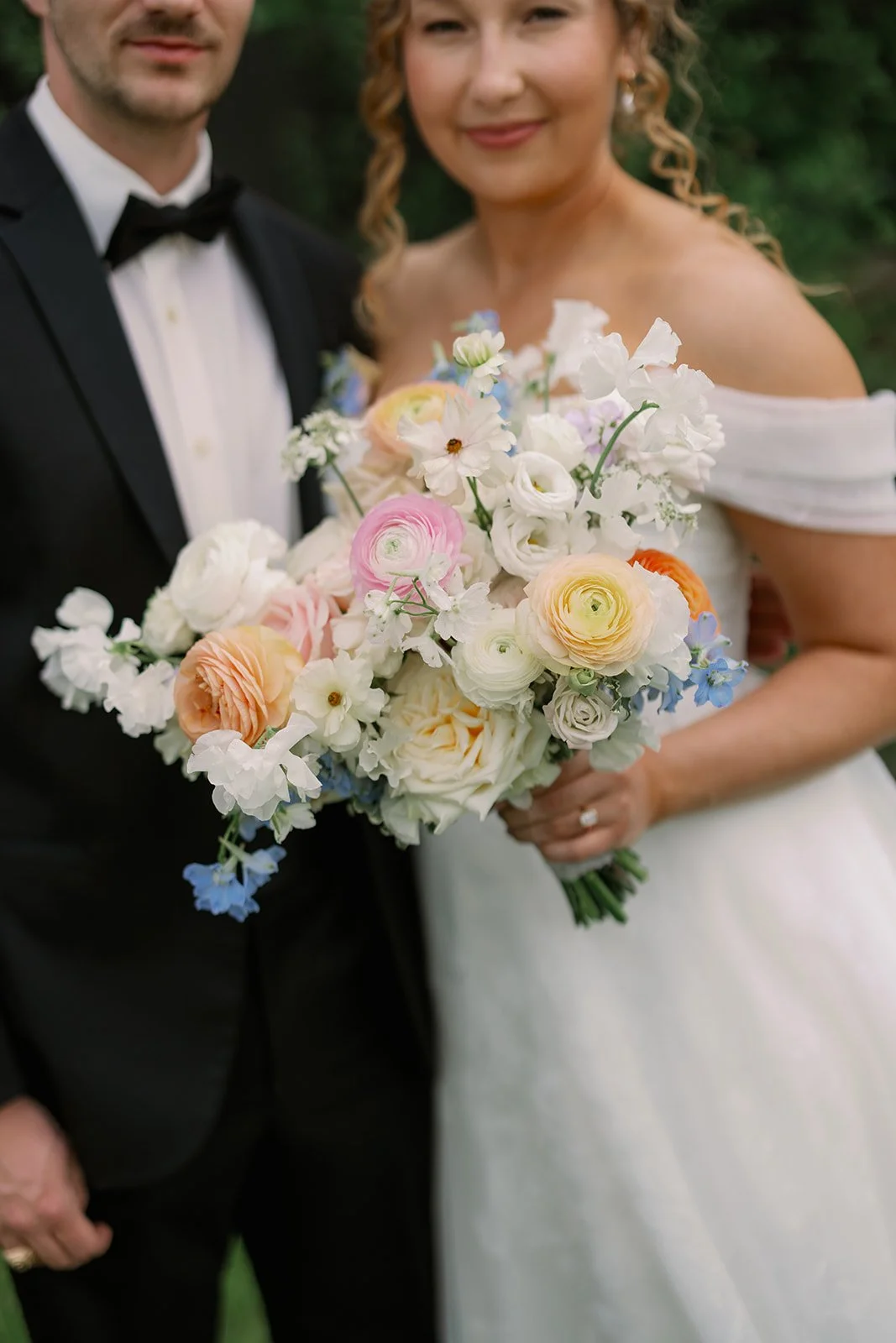 Close-up of a bride and groom holding a colorful bouquet of flowers at a wedding. The bride is wearing an off-the-shoulder white dress, and the groom is in a black tuxedo with a bow tie.