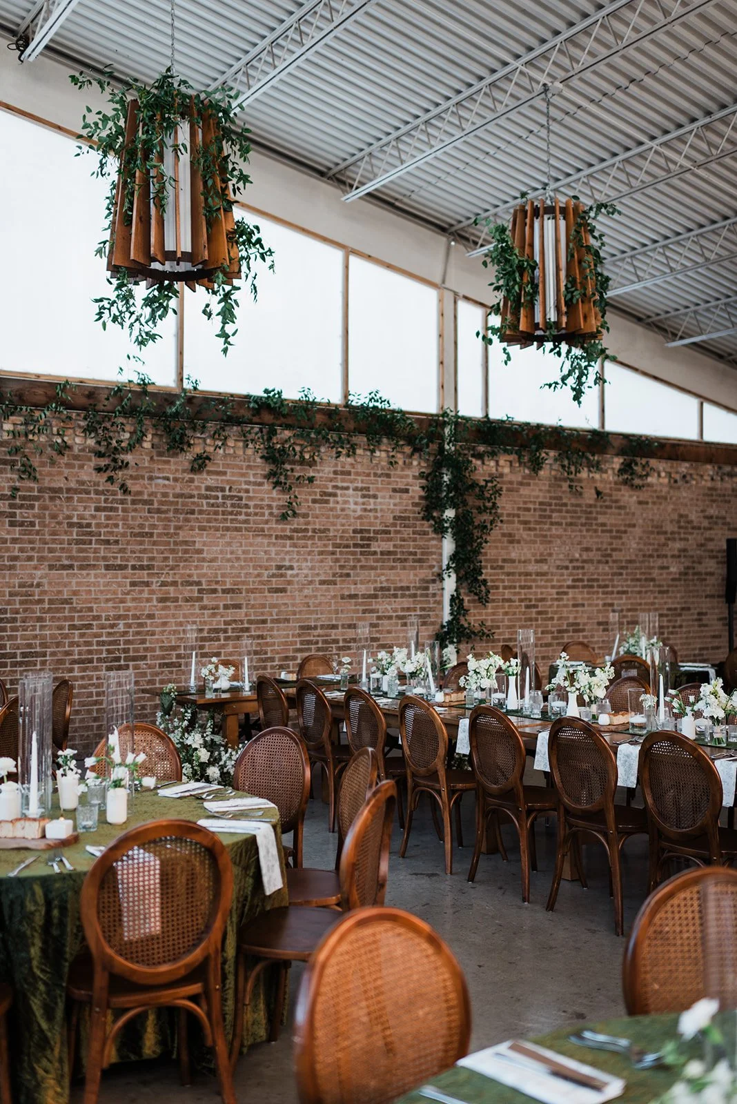 Elegant indoor dining area with long tables, wooden chairs, floral centerpieces, and hanging wooden and greenery chandeliers in a rustic venue with brick walls and a high metal ceiling.
