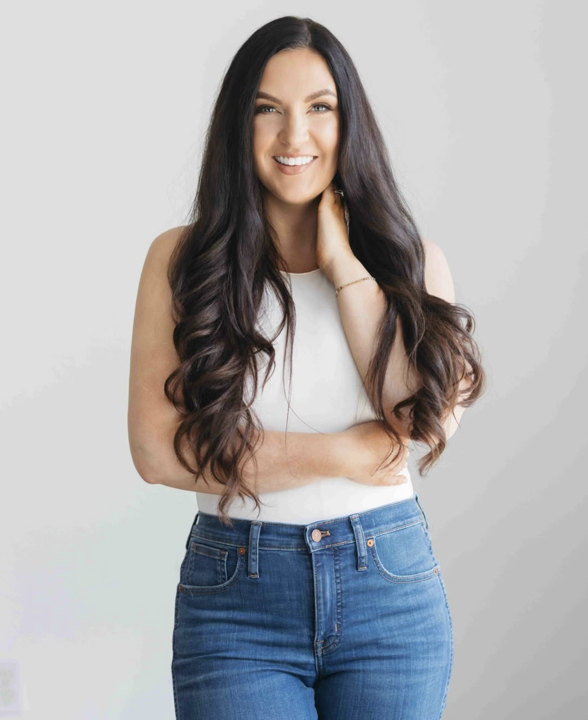 A woman with long, wavy dark brown hair and light skin, smiling, wearing a sleeveless white top and high-waisted blue jeans, standing against a plain light gray background.