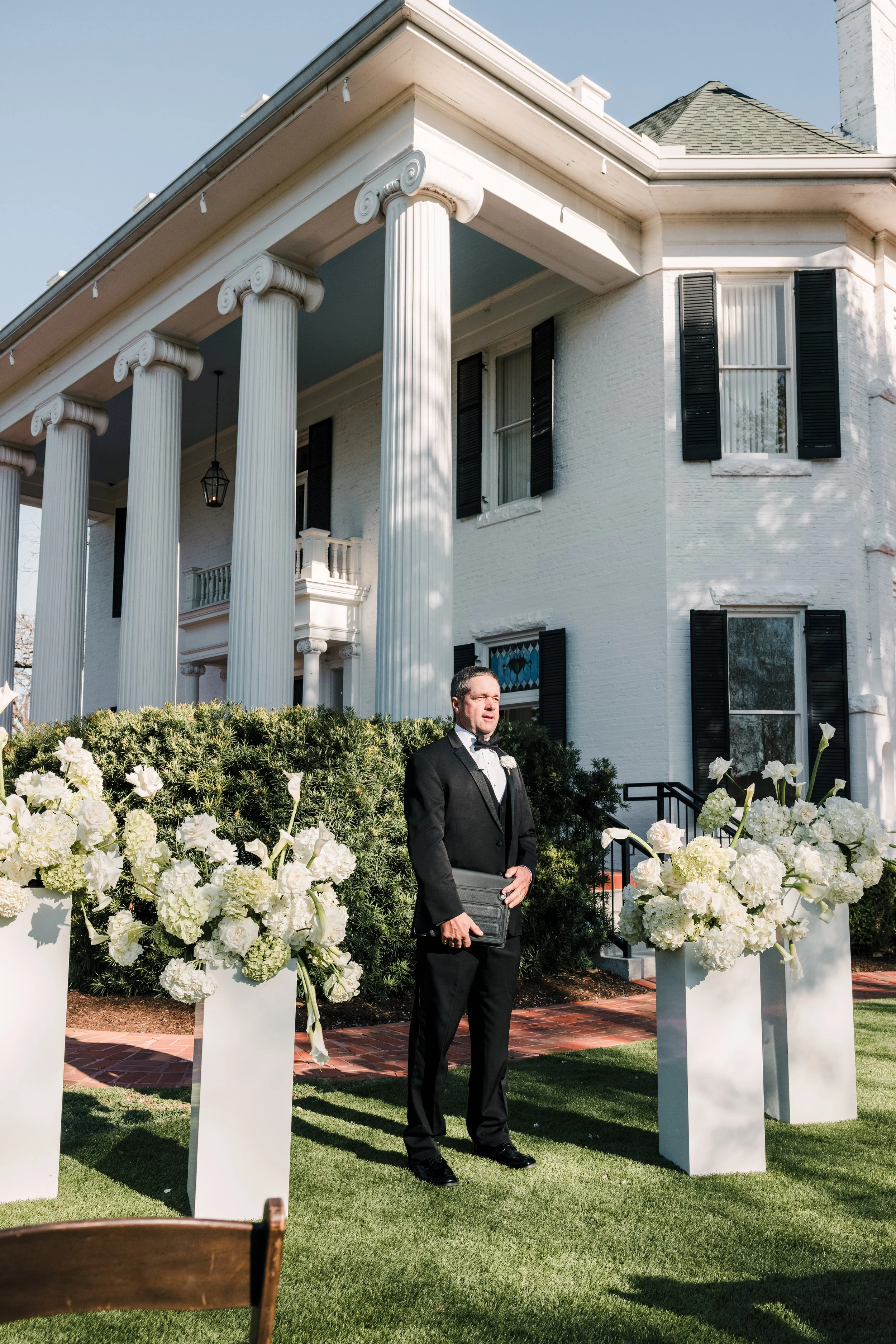 A man in formal attire stands outdoors near white flower arrangements and tall white pillars, in front of a white house with black shutters and a porch, during daylight.