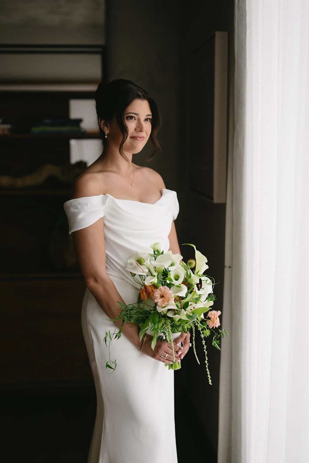 A bride in a white off-shoulder wedding gown holding a bouquet of white and pink flowers, standing by a sheer curtain.