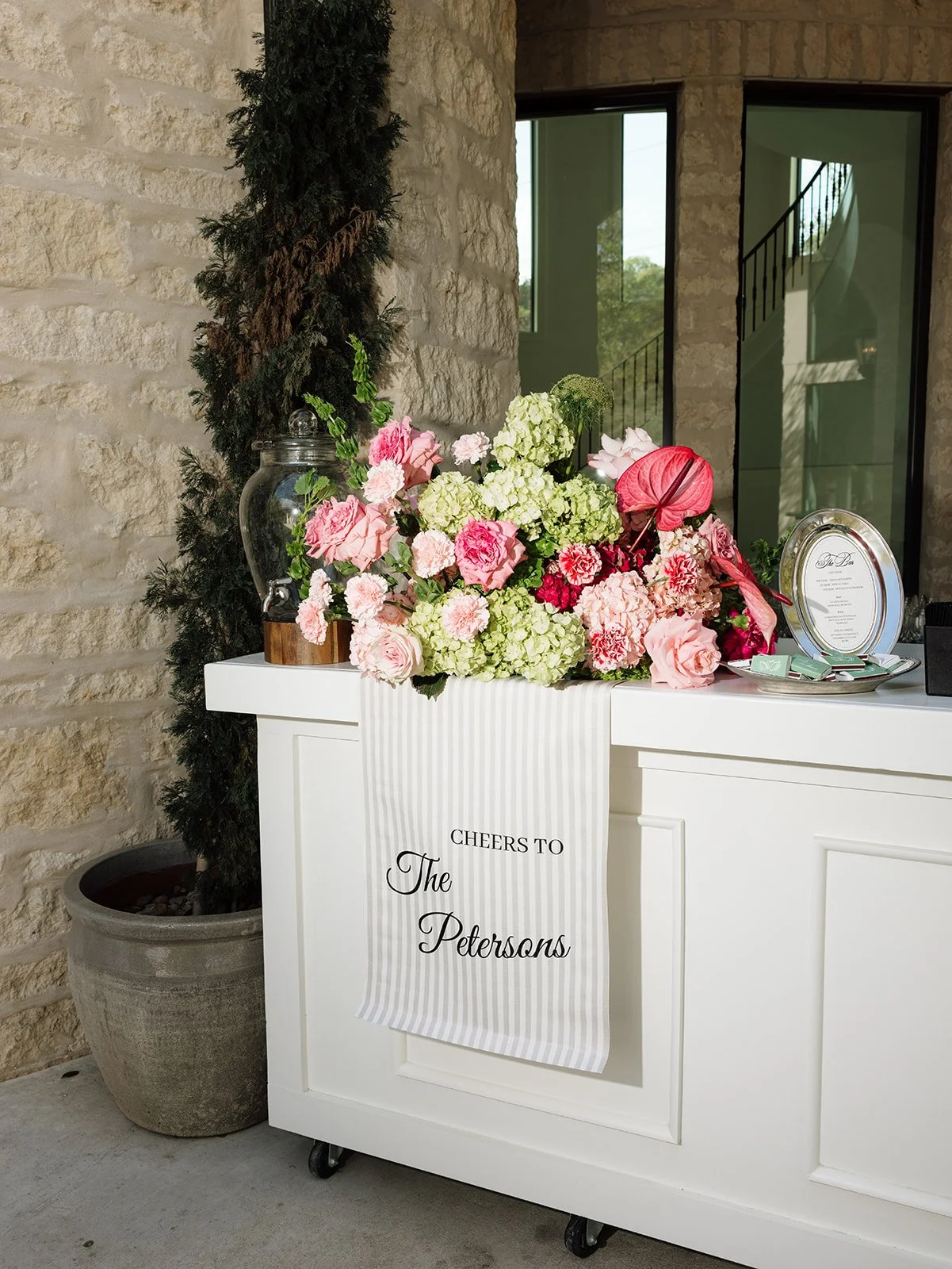 A white rolling cart with a large arrangement of pink and white flowers, a framed menu, and a glass jar with a spigot. A sign on the front of the cart says 'Cheers to The Petersons.' There is a potted evergreen tree and a stone wall in the background