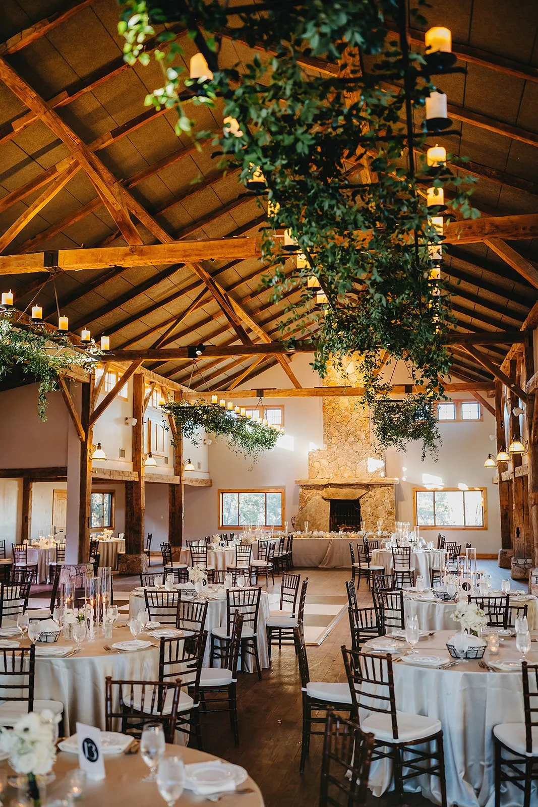 A rustic indoor event space decorated with hanging greenery and candles, featuring round tables with white tablecloths and black chairs, a stone fireplace, and natural light from large windows.