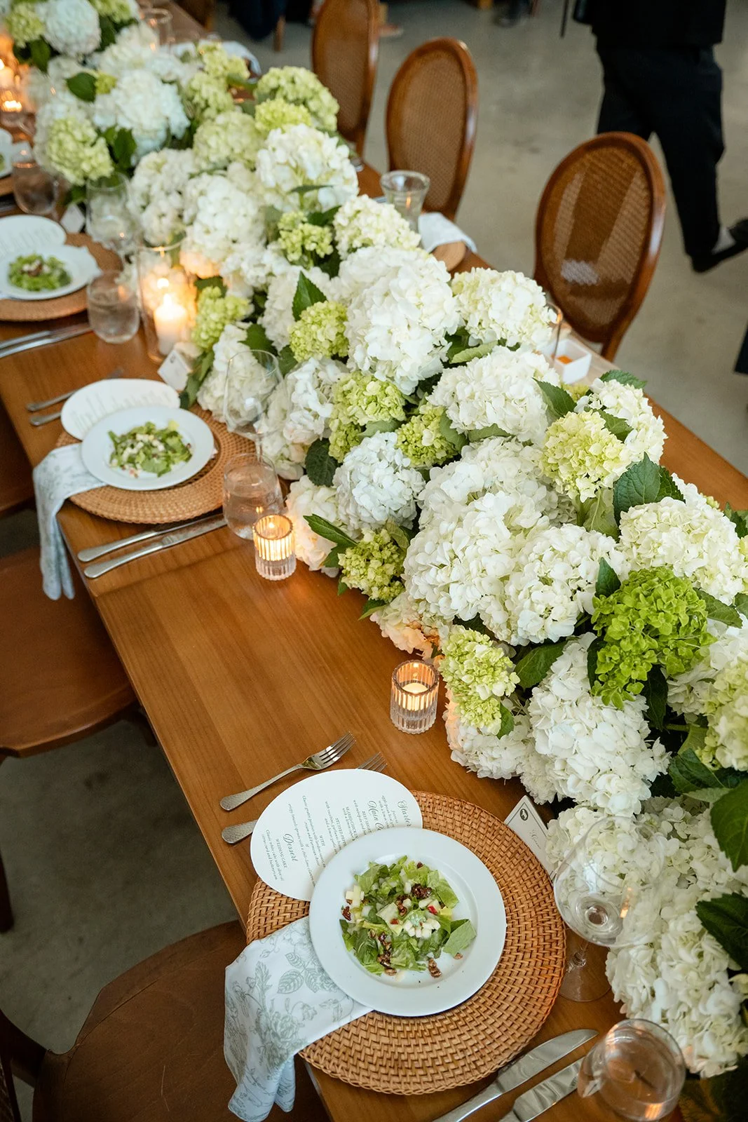 A long dining table decorated with a lush centerpiece of white and green hydrangeas, surrounded by wooden chairs with woven backs. The table is set with white plates, silverware, water glasses, candles, and printed menus. Small salads are served on e